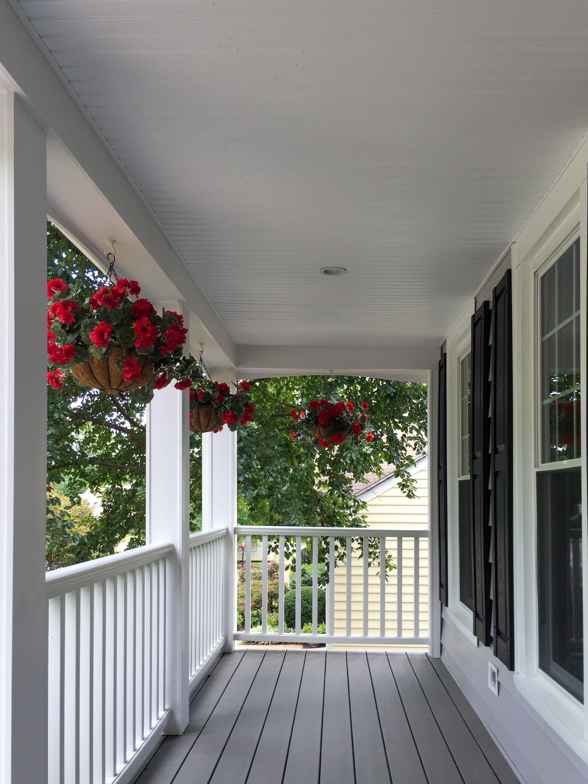 A covered, wooden porch with grey floorboards, white railings, and three hanging baskets of vibrant red flowers.