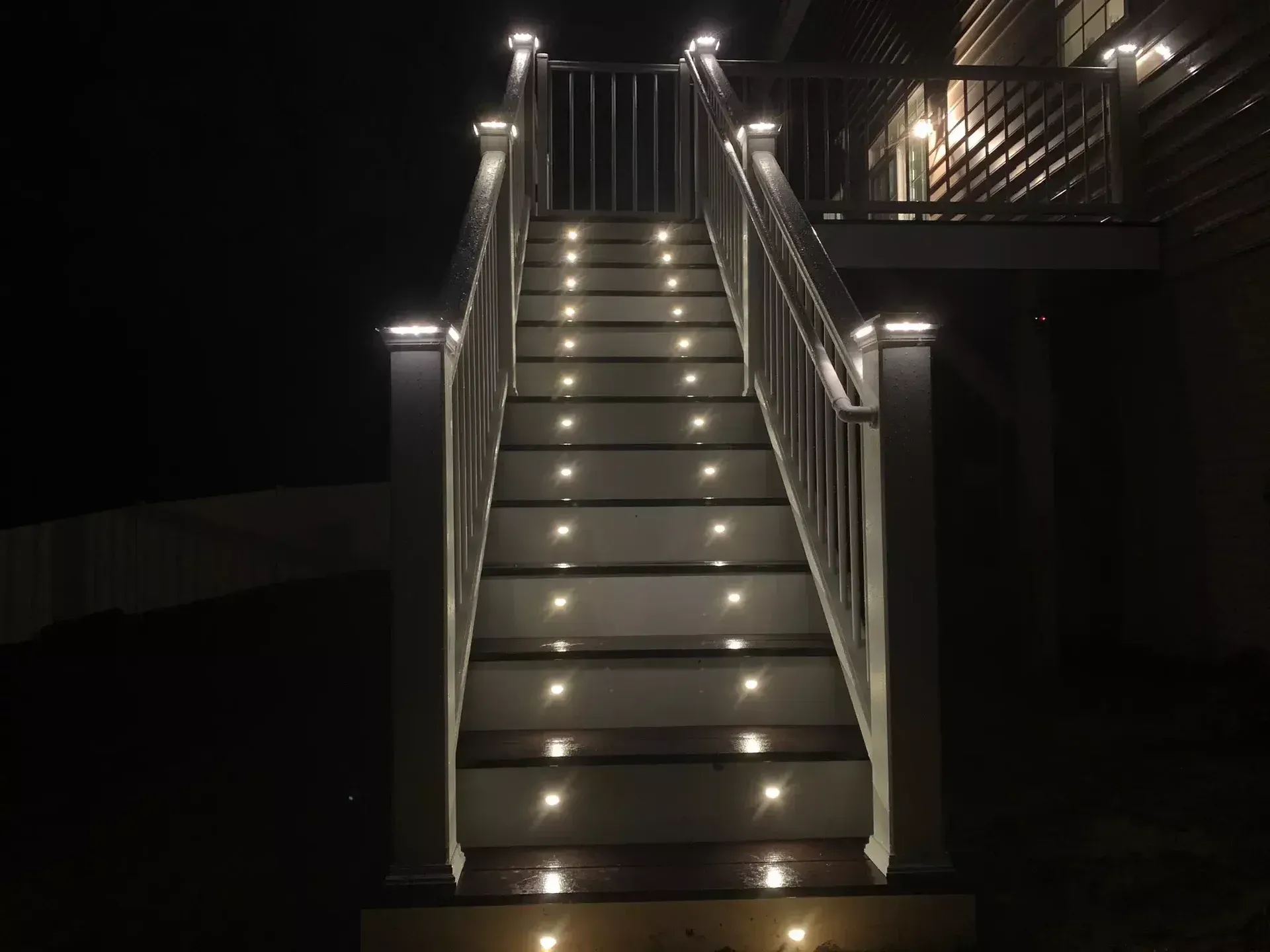 A set of outdoor wooden stairs at night, illuminated by recessed lights on each step and post caps on the railing.