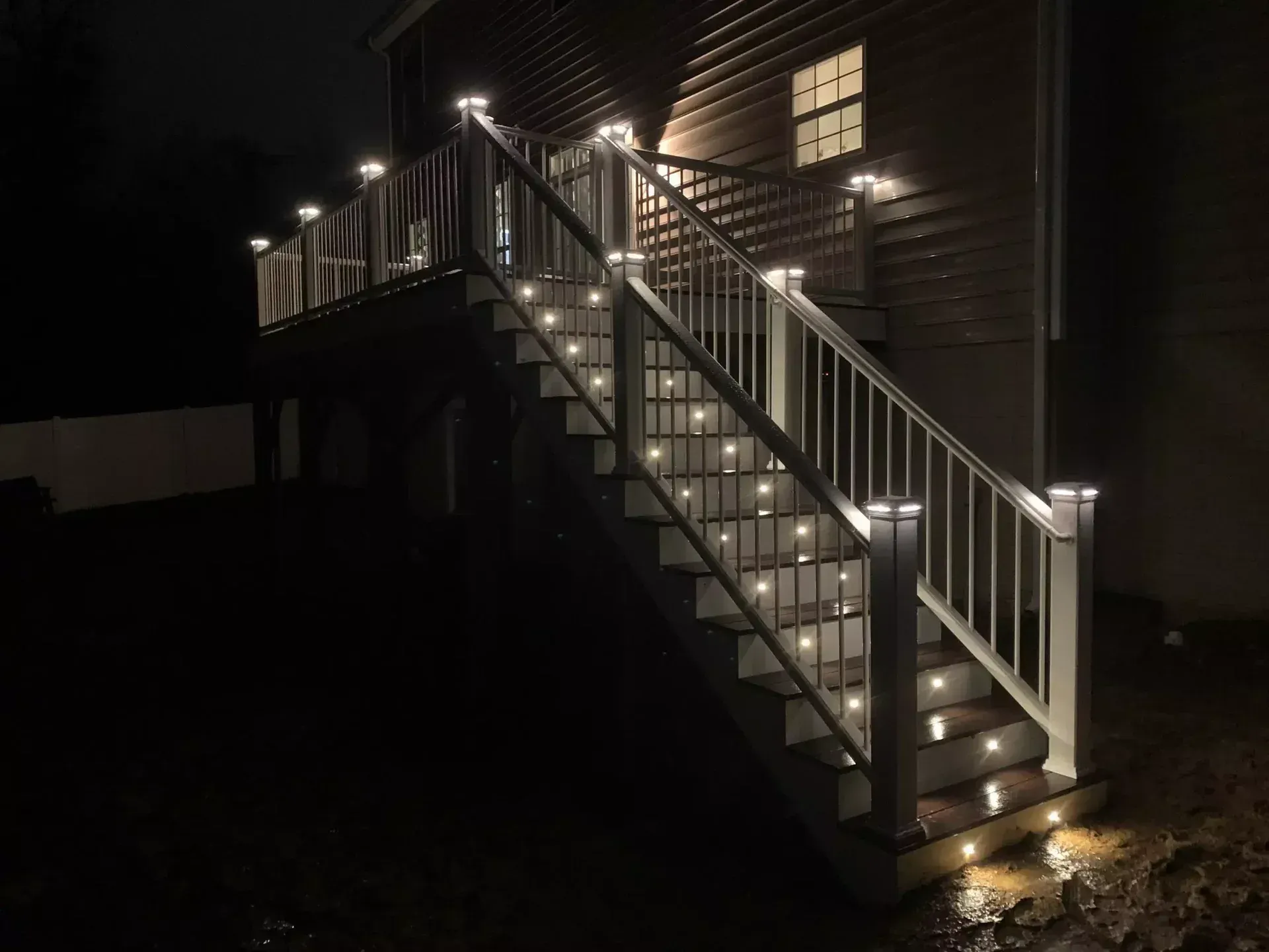 A wooden staircase leading up to a deck at night, illuminated by soft white LED lights on the posts and each step.