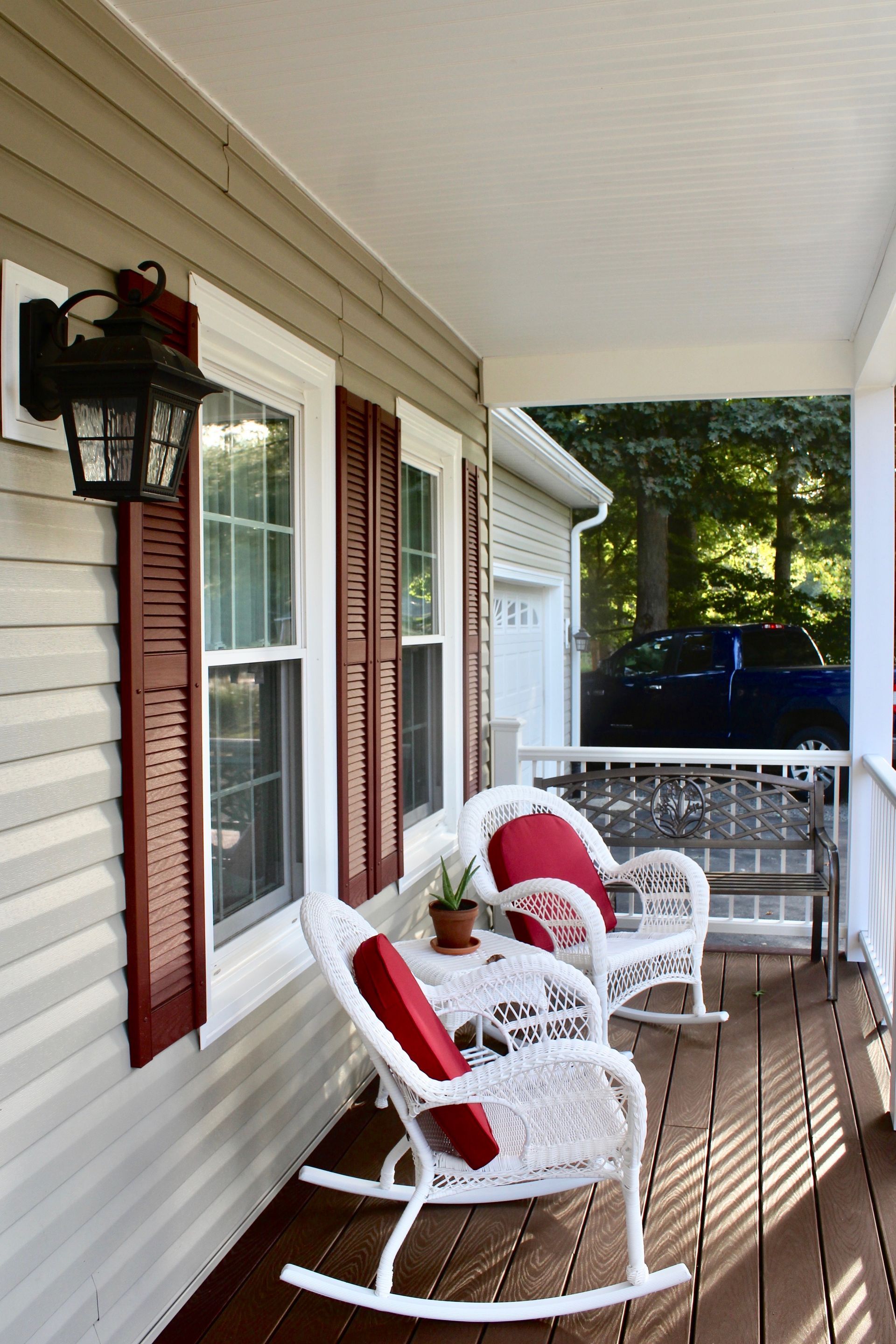 Two white wicker rocking chairs with red cushions on a wooden porch, featuring red shutters and a black wall lantern.