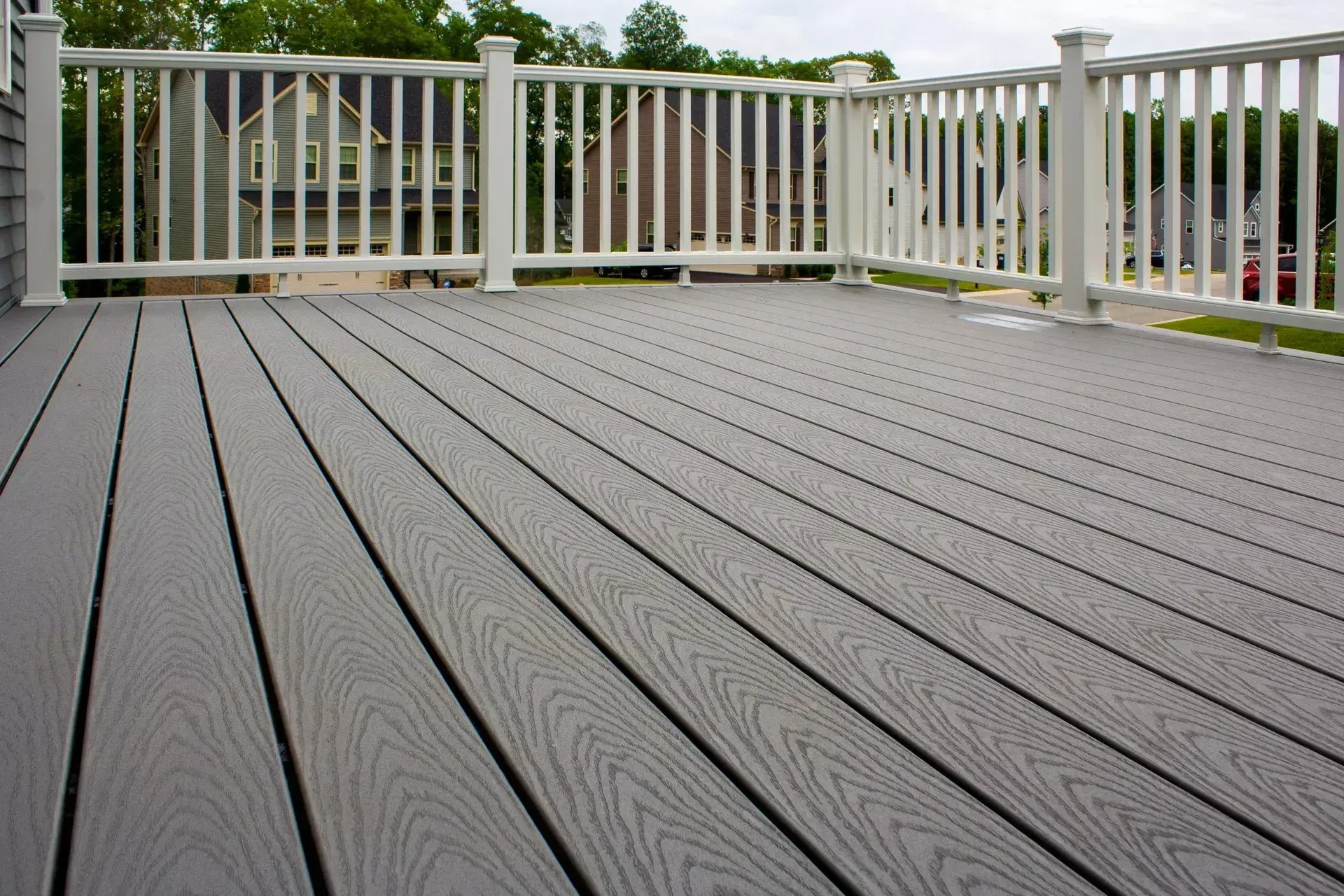 A grey wood-composite deck with white railings overlooks a house and trees in a residential backyard.