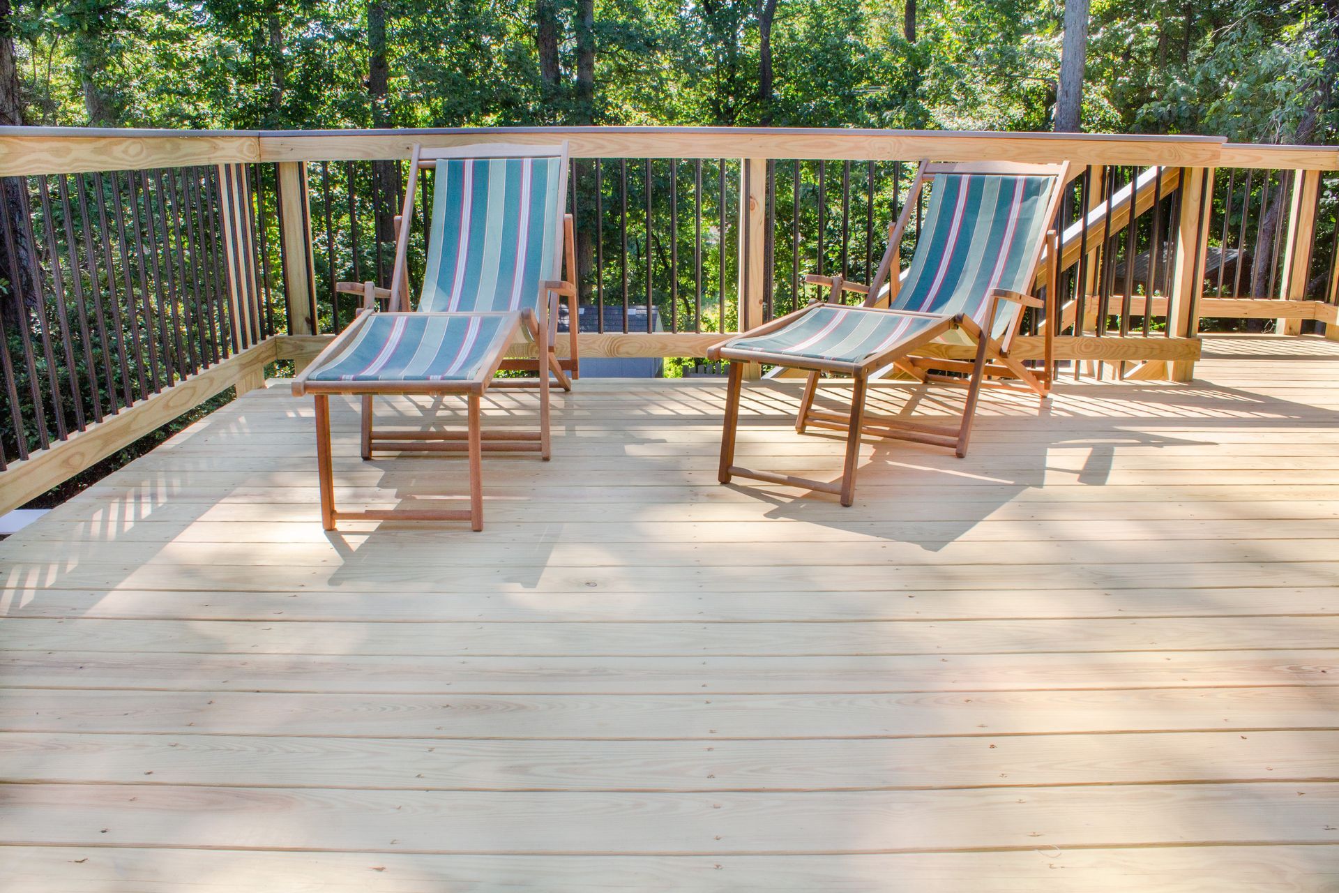 Two striped sling chairs with matching footrests sit on a sunlit wooden deck overlooking a leafy green forest.