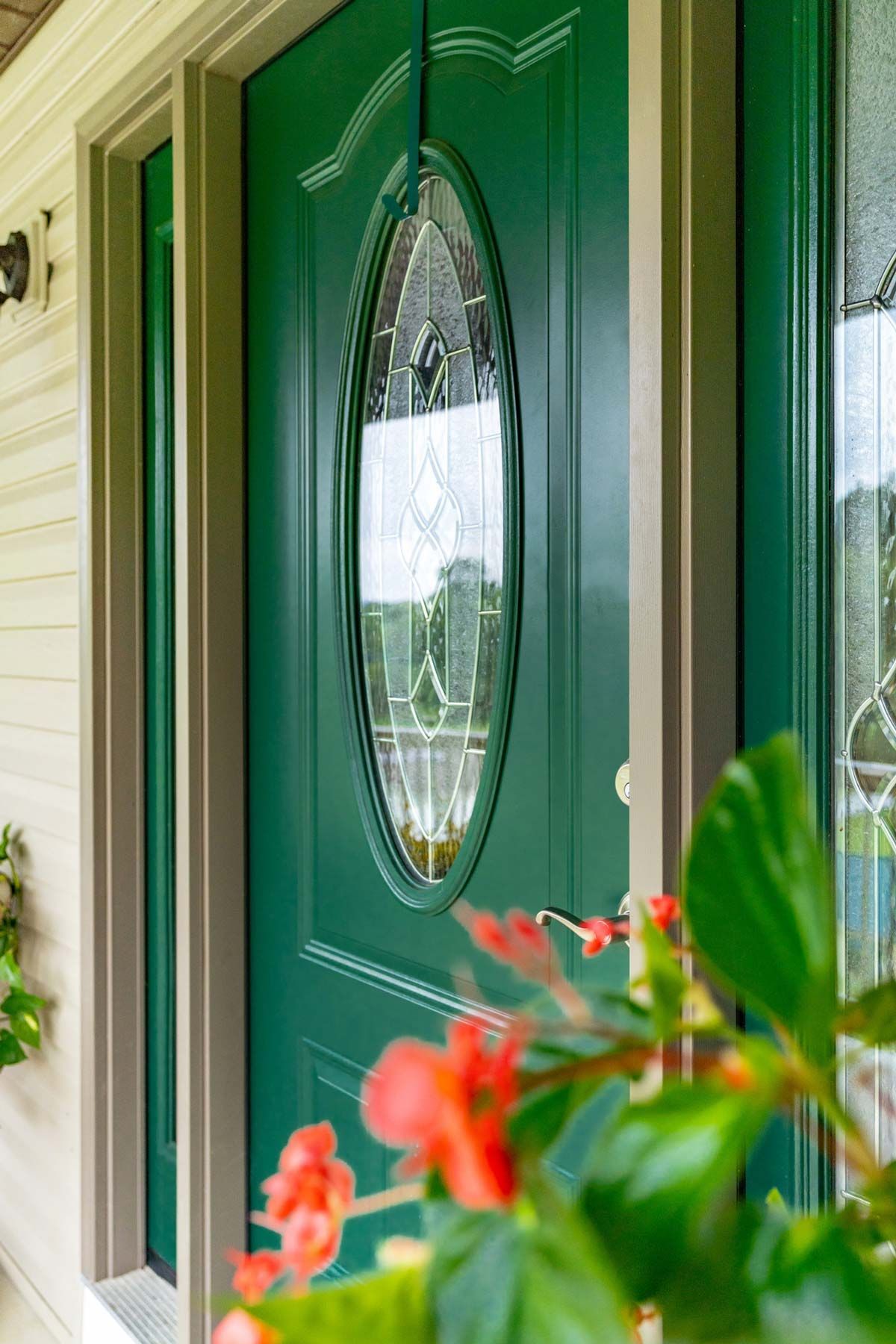 A green front door with an oval stained-glass window, framed by light-colored siding and out-of-focus red flowers nearby.