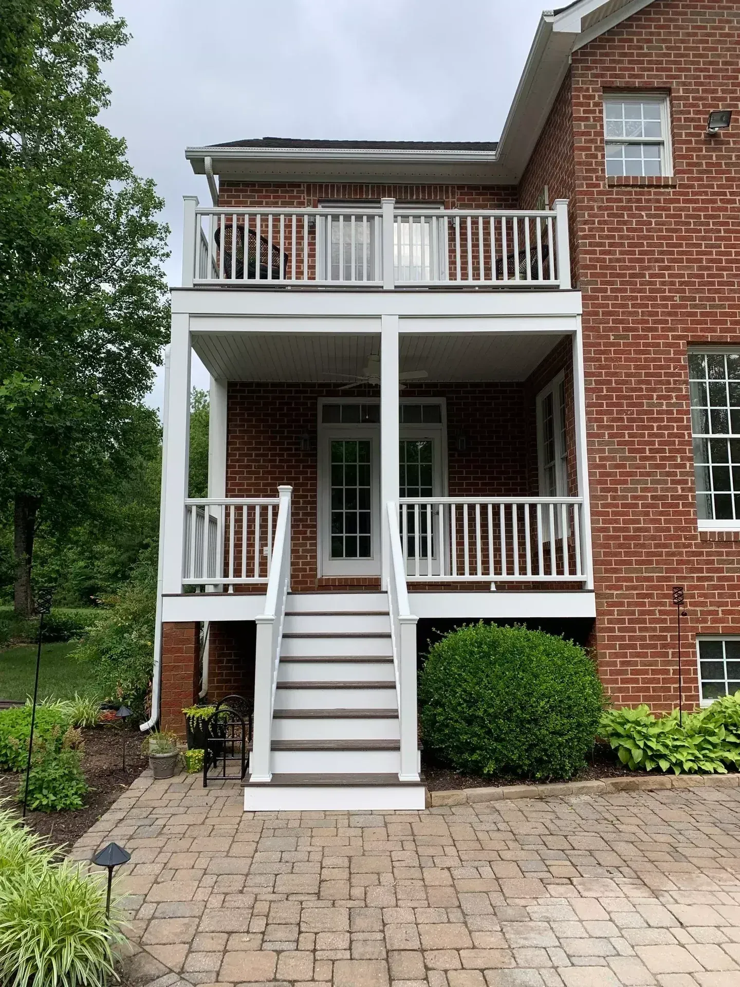 A two-story brick home with a white-railed deck on the upper level and a porch with stairs leading to a brick patio.