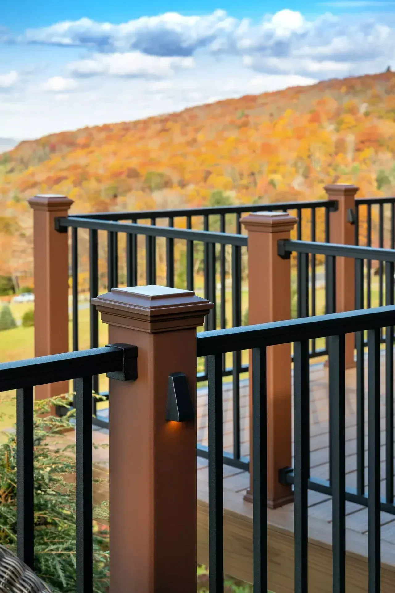 A brown deck railing with black metal spindles and integrated post lights, overlooking a mountain with autumn foliage.