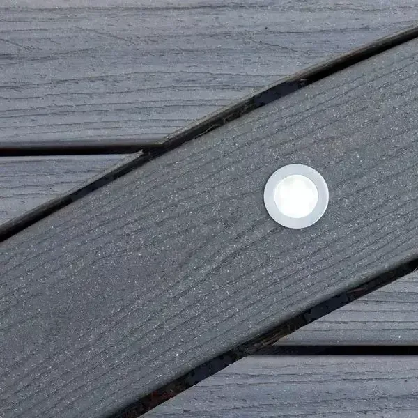A circular LED deck light embedded flush into a dark gray, wood-grain textured composite deck plank.
