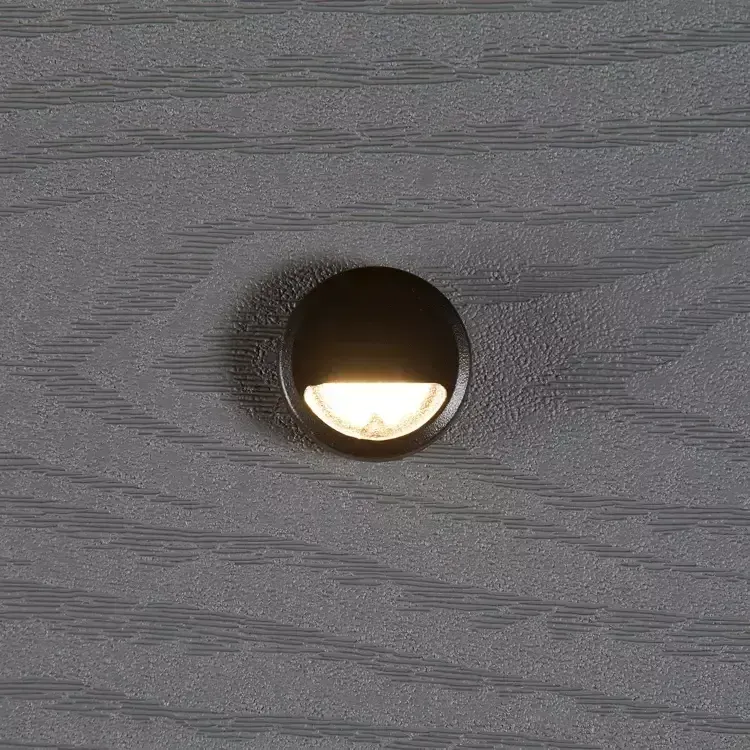 A small, circular black recessed light with a warm, downward-facing glow, installed on a gray wood-grain surface.
