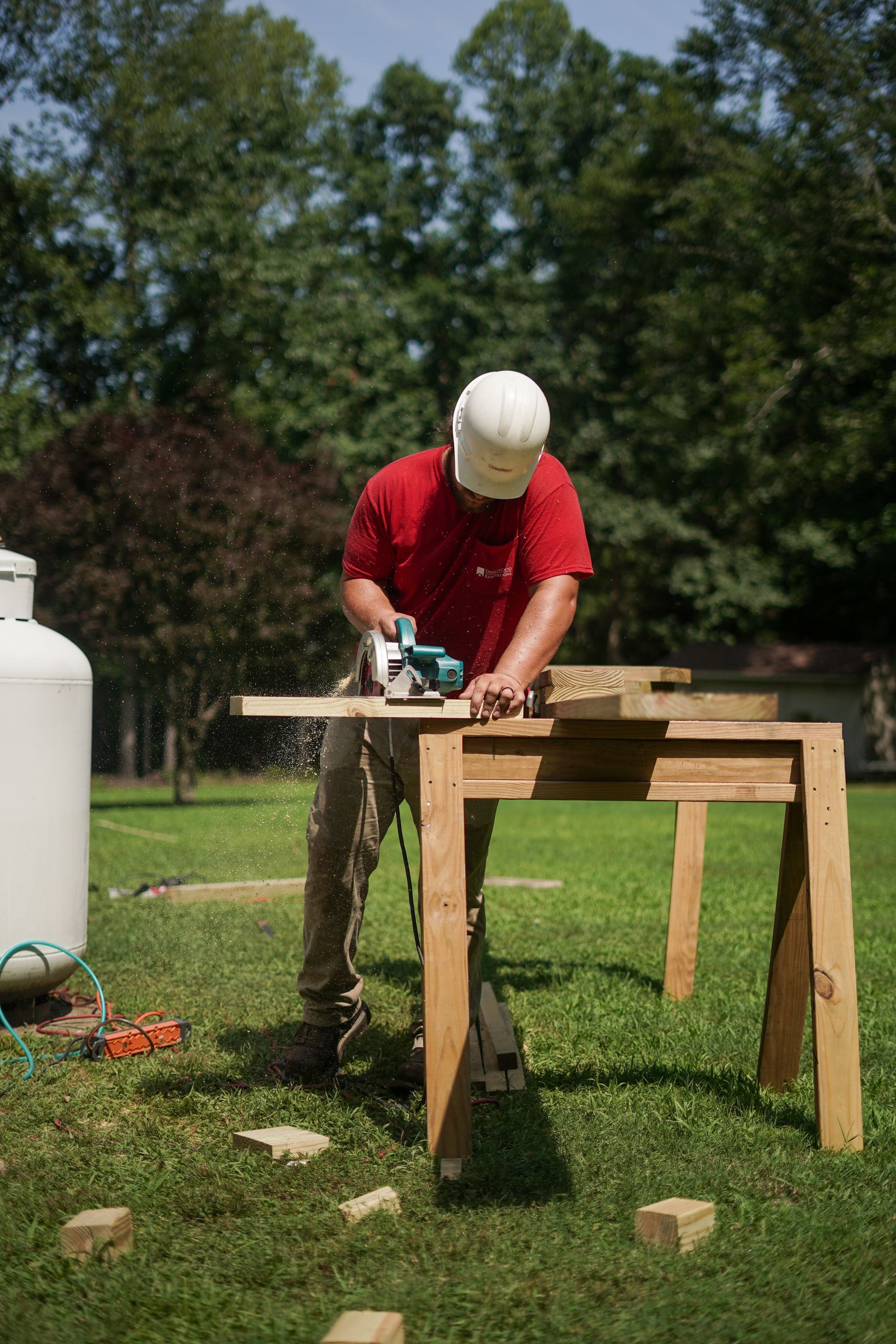 Person in a white hard hat using a power saw on a wooden workbench outdoors.