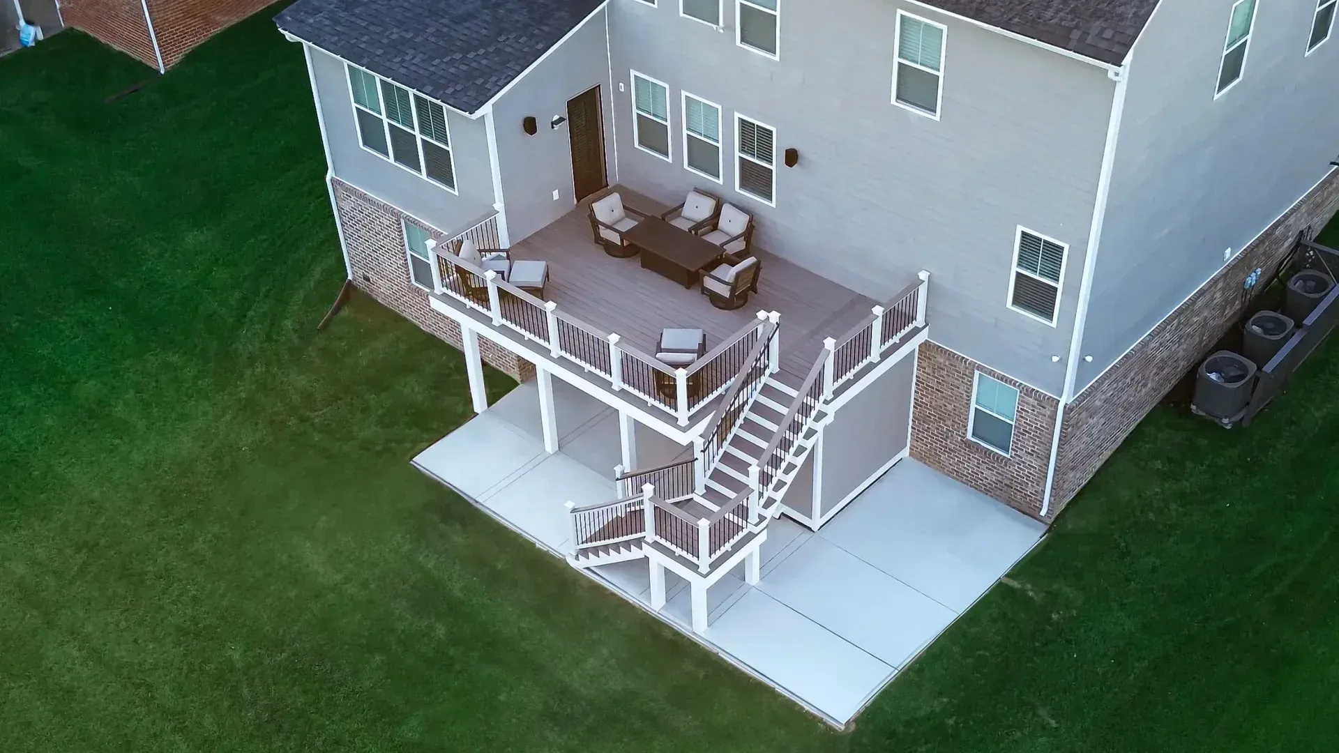 Aerial view of a gray house with a wooden deck featuring patio furniture, stairs to a concrete patio, and a grassy yard.