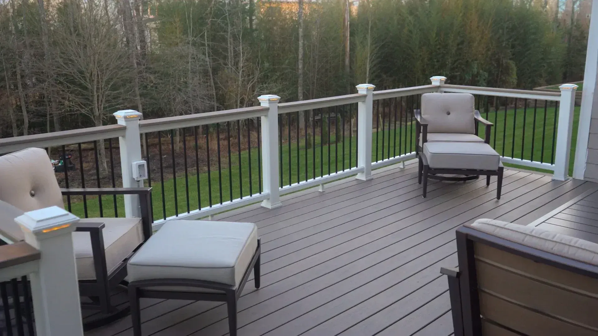 Outdoor deck with two armchairs and matching ottomans, overlooking a backyard and treeline at sunset.