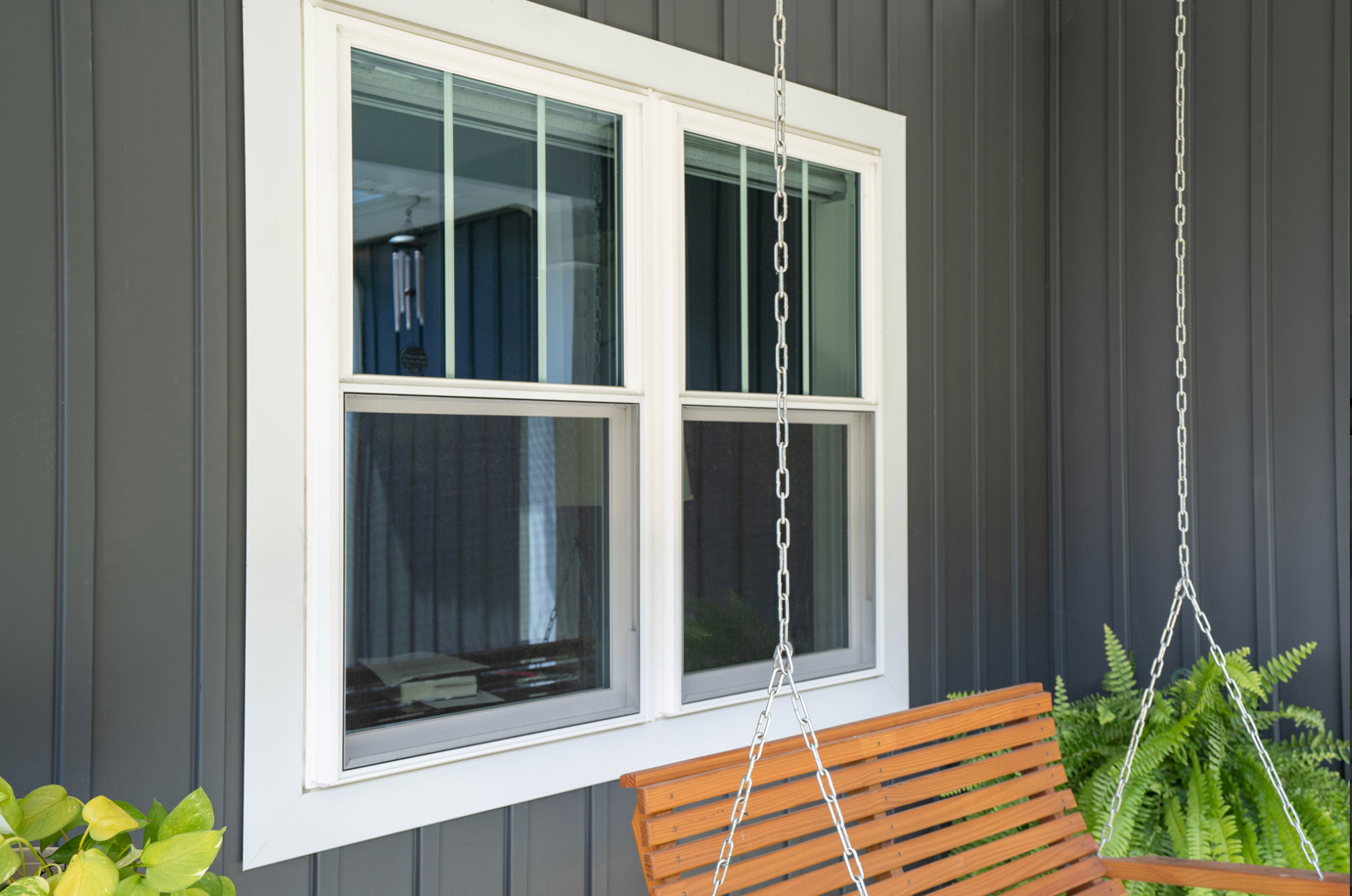 Two wicker armchairs with white cushions sit on a porch in front of a dark-framed double window and hanging greenery.