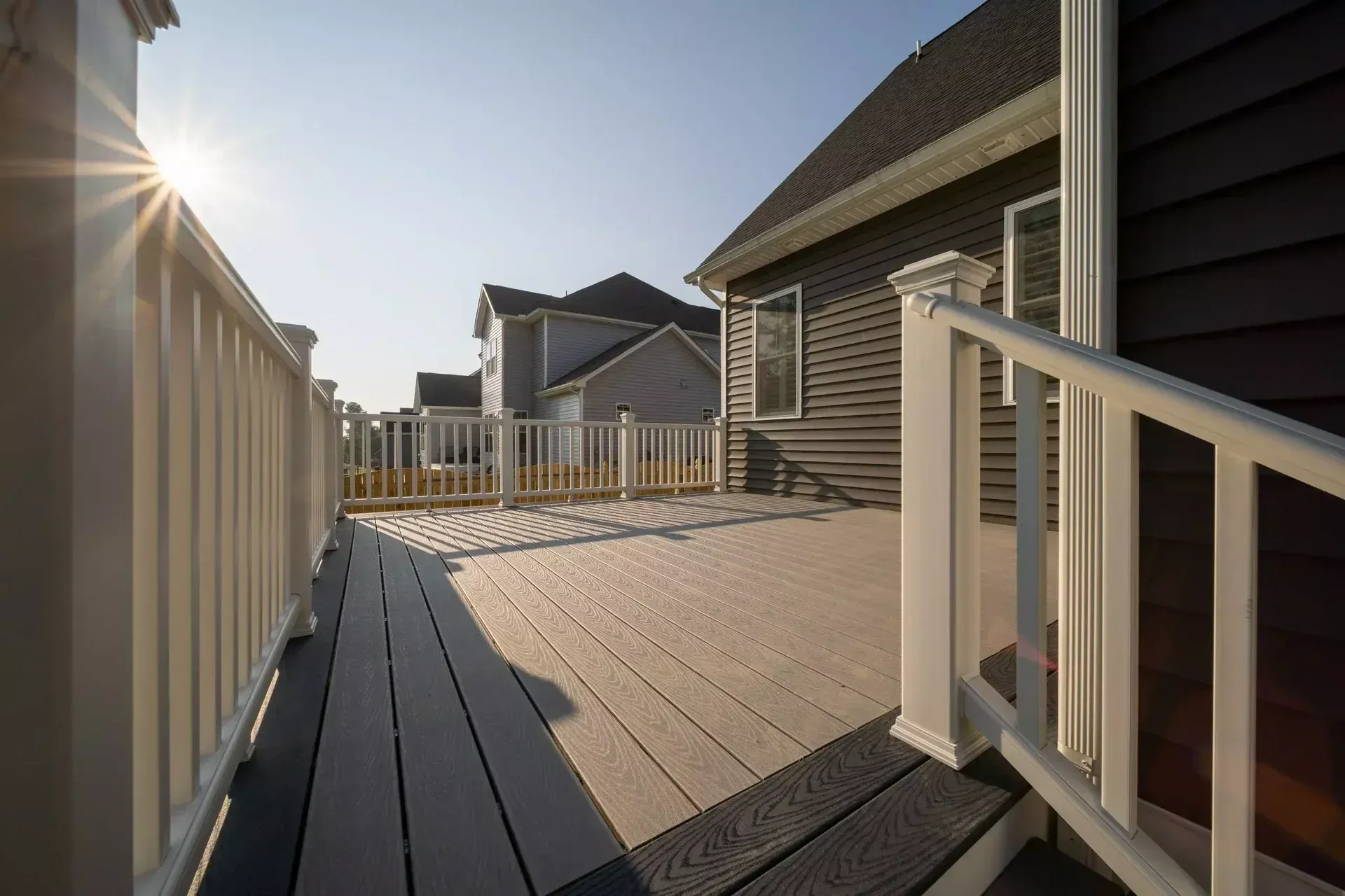 A sunny deck with light-colored planks and dark border boards, white railings, and a view of neighboring houses.