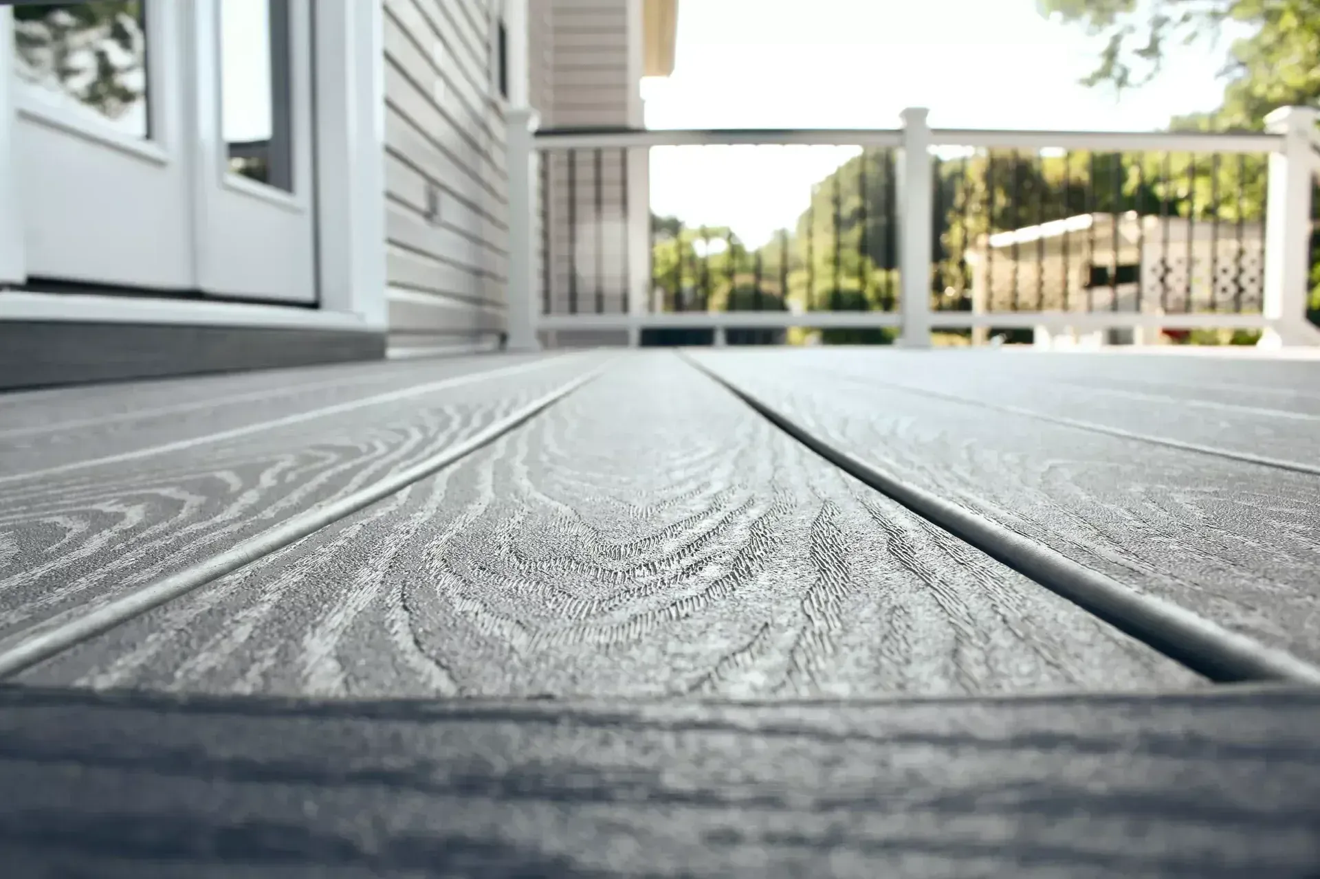 Low-angle, close-up view of grey, wood-grain textured composite deck boards leading toward a house exterior and railing.
