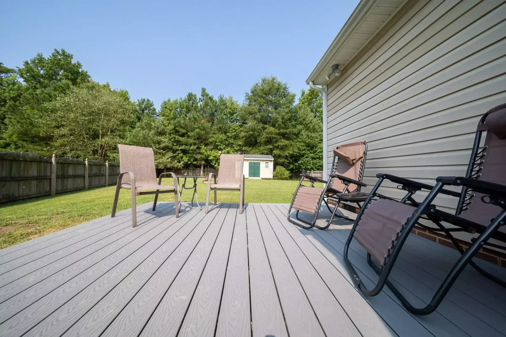 A gray backyard deck featuring two lounge chairs in the foreground and two stationary chairs facing a wooded yard.