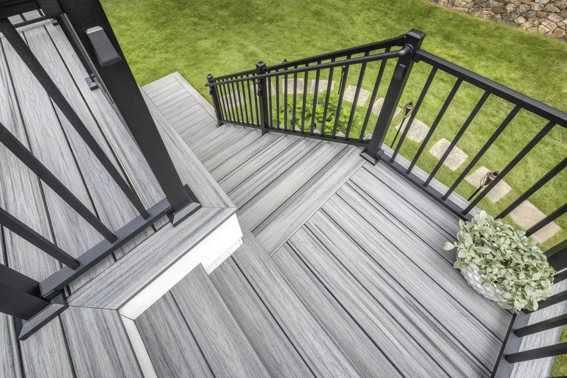 Gray wood-look composite deck stairs with black metal railings lead down to a grassy backyard with stone pavers.