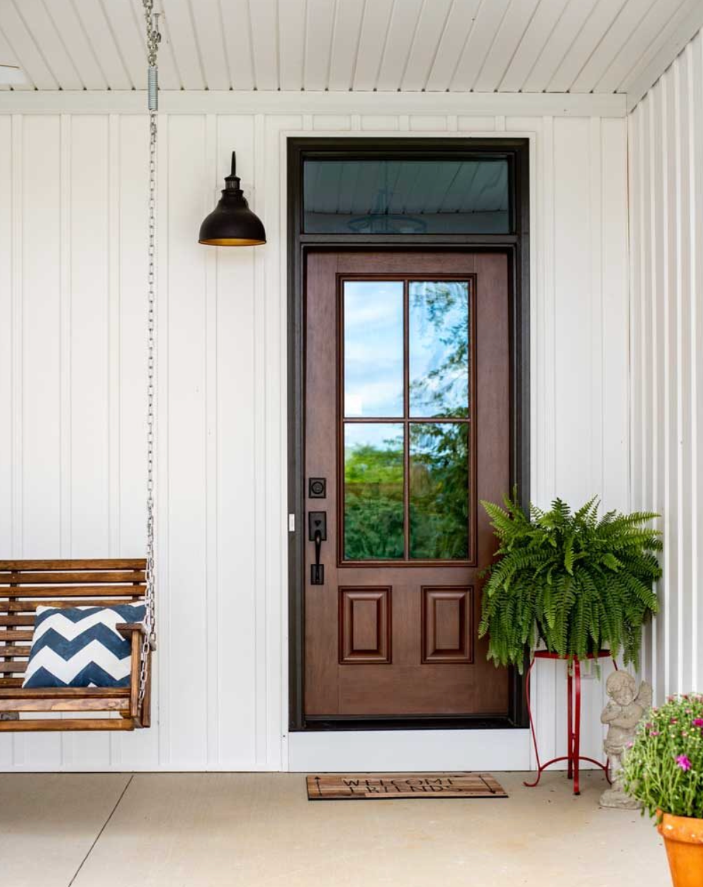 Front porch with brown door, hanging black light, swing bench, potted fern, and colorful rug.
