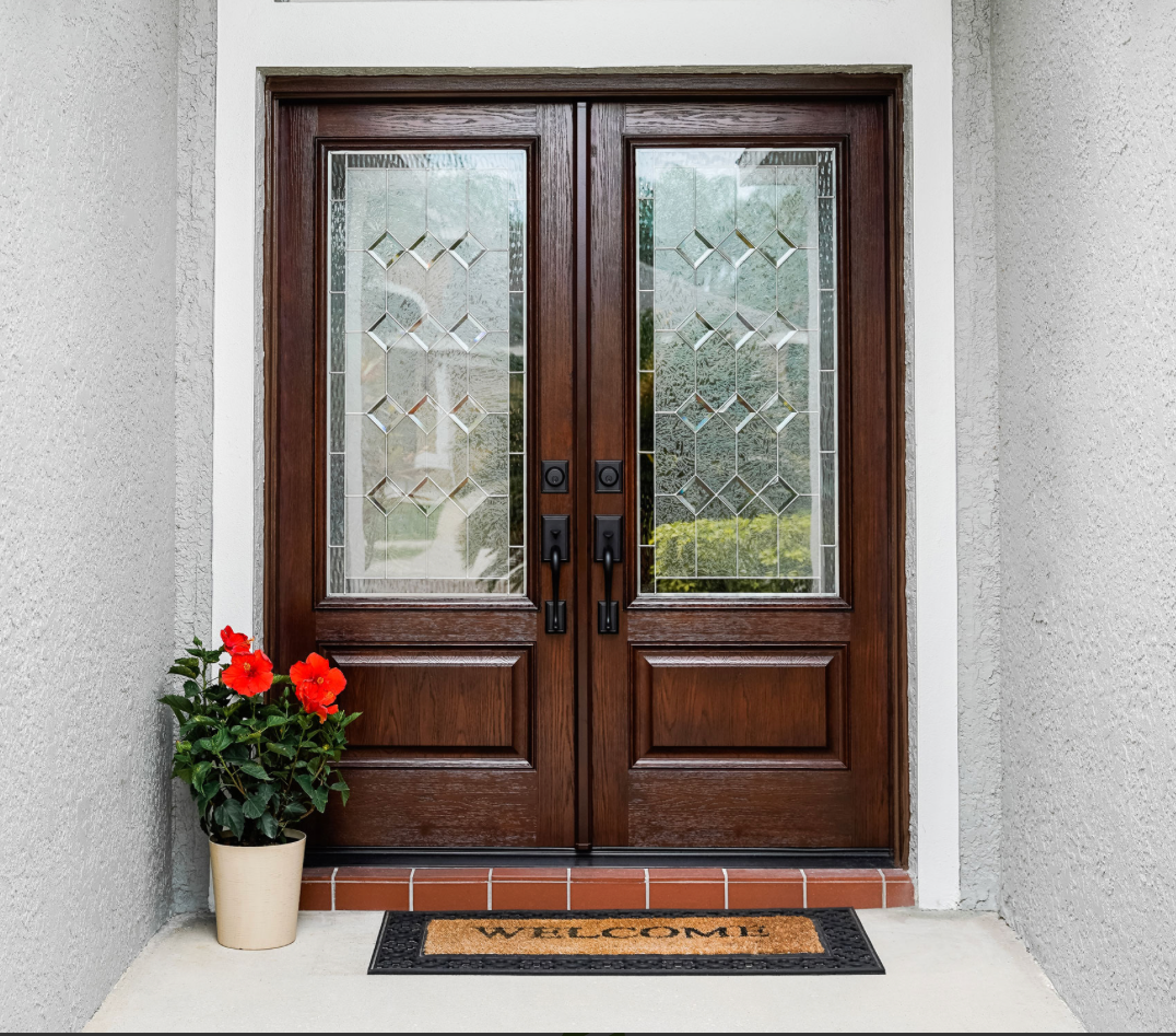 Dark wooden double doors with decorative glass panels, an arched window above, and a potted red hibiscus on the porch.