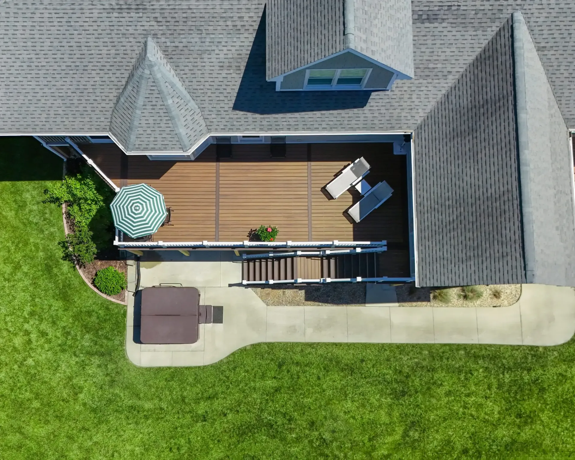 An aerial view of a backyard with a wooden deck featuring lounge chairs and an umbrella, next to a patio with a hot tub.