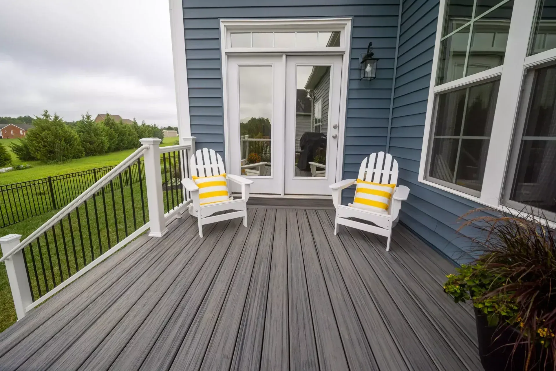 Two white Adirondack chairs with yellow-striped pillows sit on a gray deck outside a blue house with white trim.