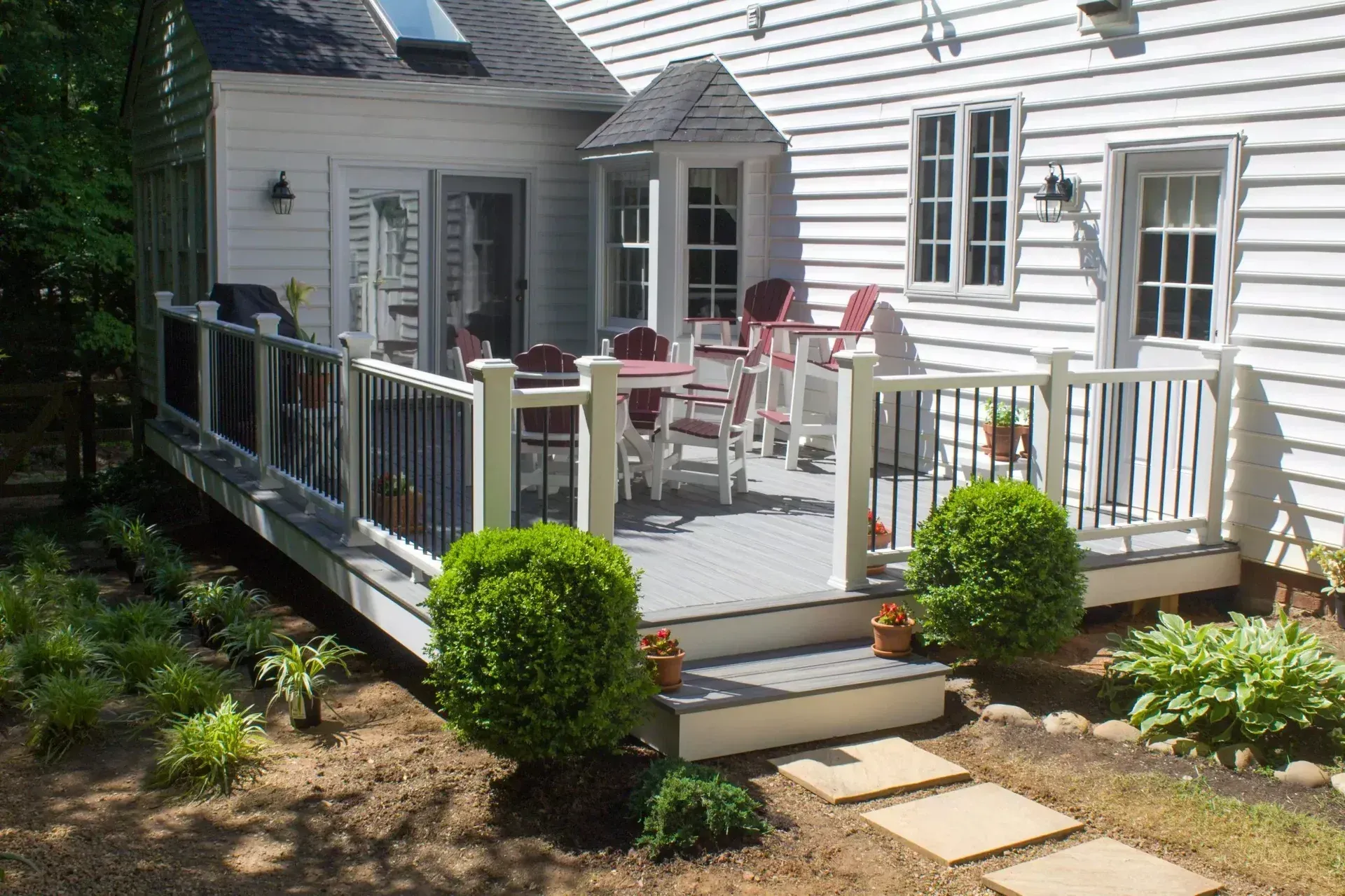 A white-sided house with a raised wooden deck featuring a dining set, white railings, and manicured shrubs in the yard.