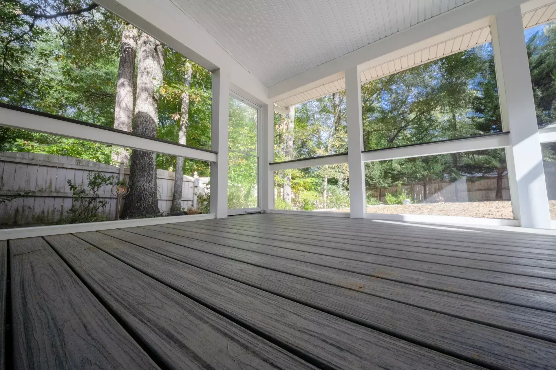 A low-angle view of a grey wooden deck inside a screened-in porch, looking out at a backyard with trees and a white fence.