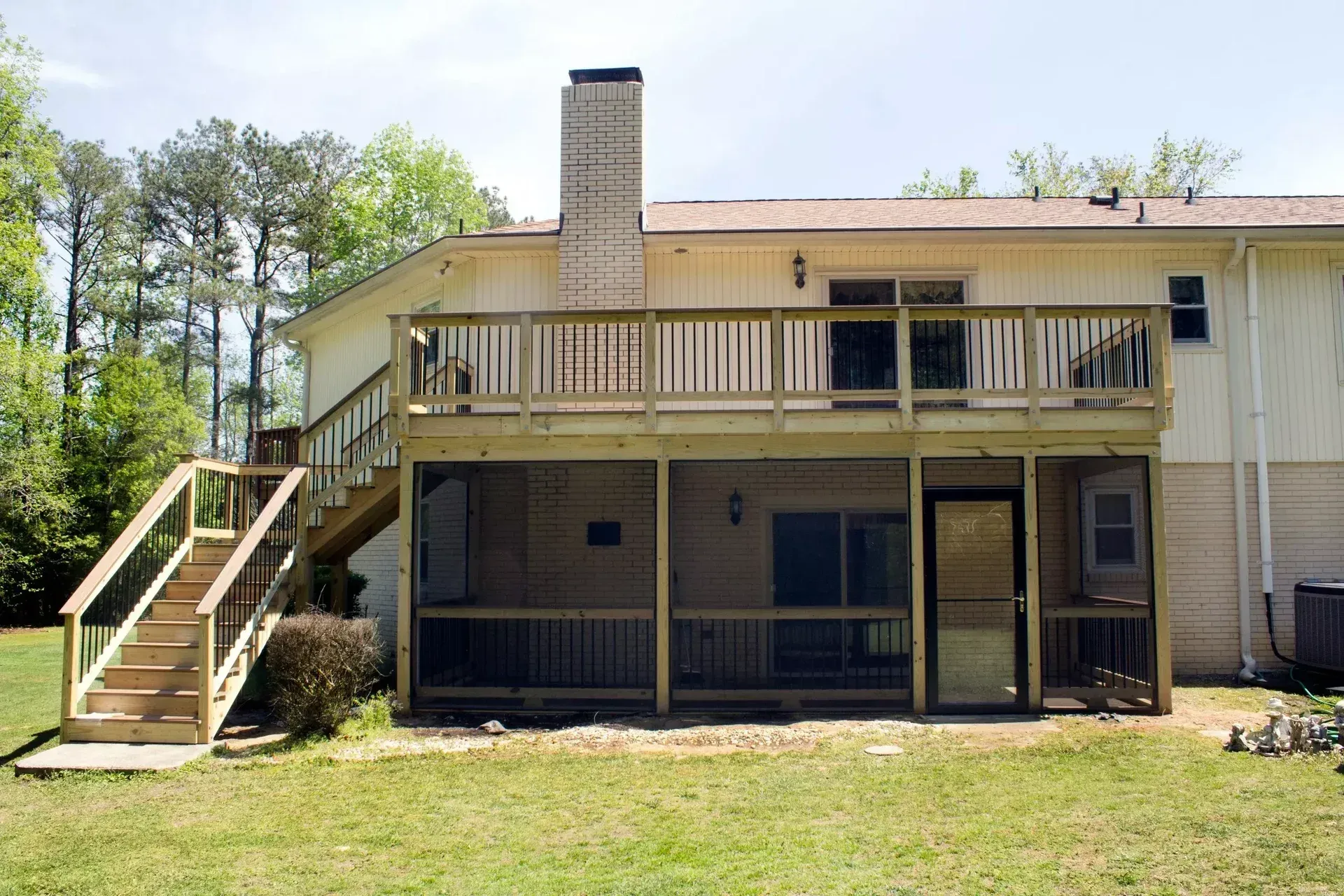 A light-colored two-story house with a wooden deck and a screened-in porch with stairs leading to the lawn.