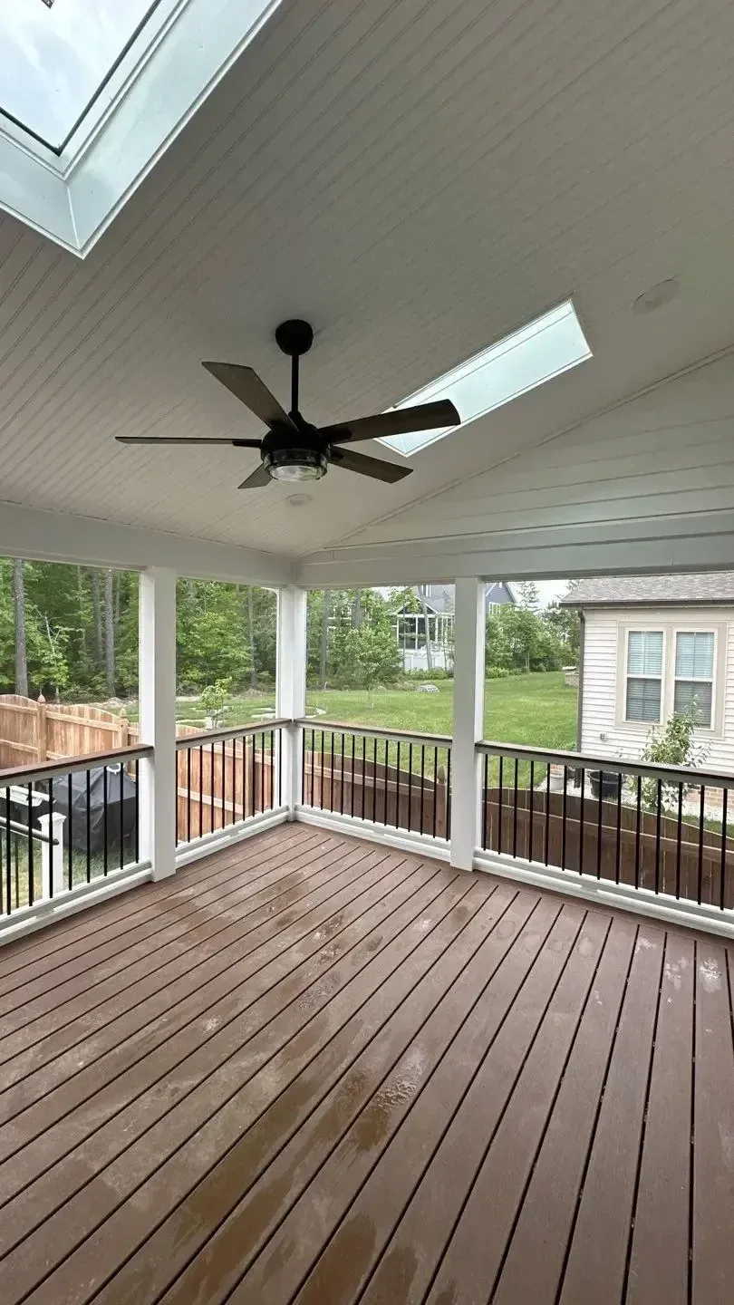 A covered outdoor deck featuring wood-toned planks, white support columns, a black ceiling fan, and two large skylights.
