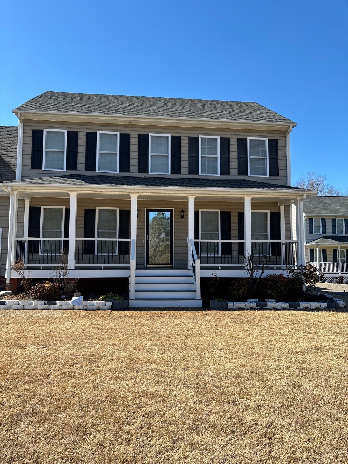 A light-colored two-story house with a white front porch, stairs, brick pillars, and landscaped garden beds.