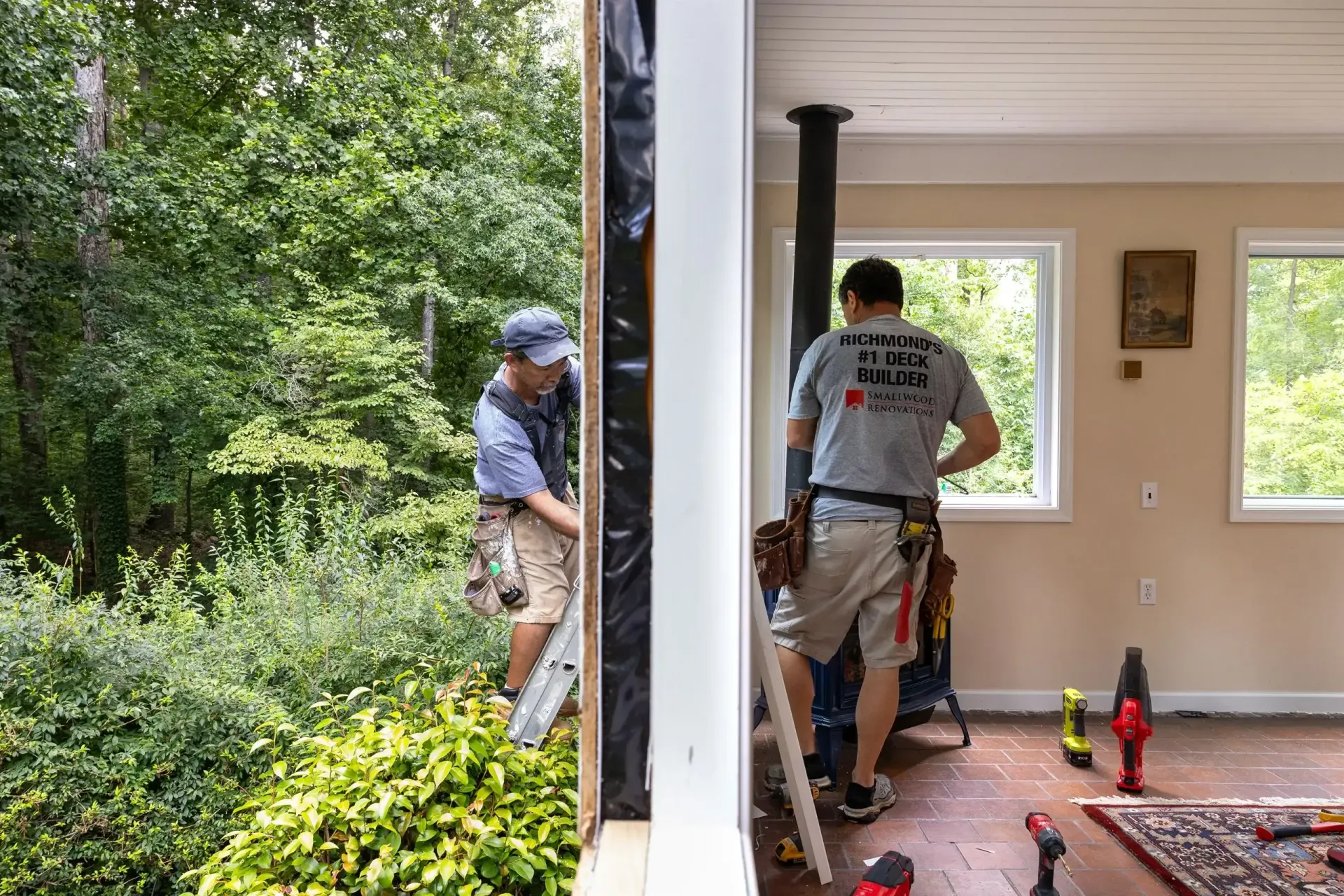 Two workers perform interior and exterior renovations on a room with large windows overlooking a wooded area.