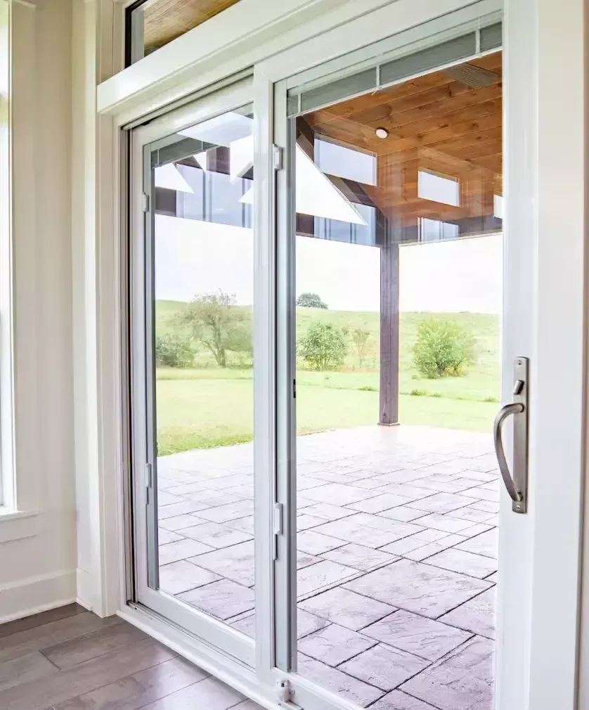 A white sliding glass door opening onto a stone patio with a view of a grassy field and a covered porch.