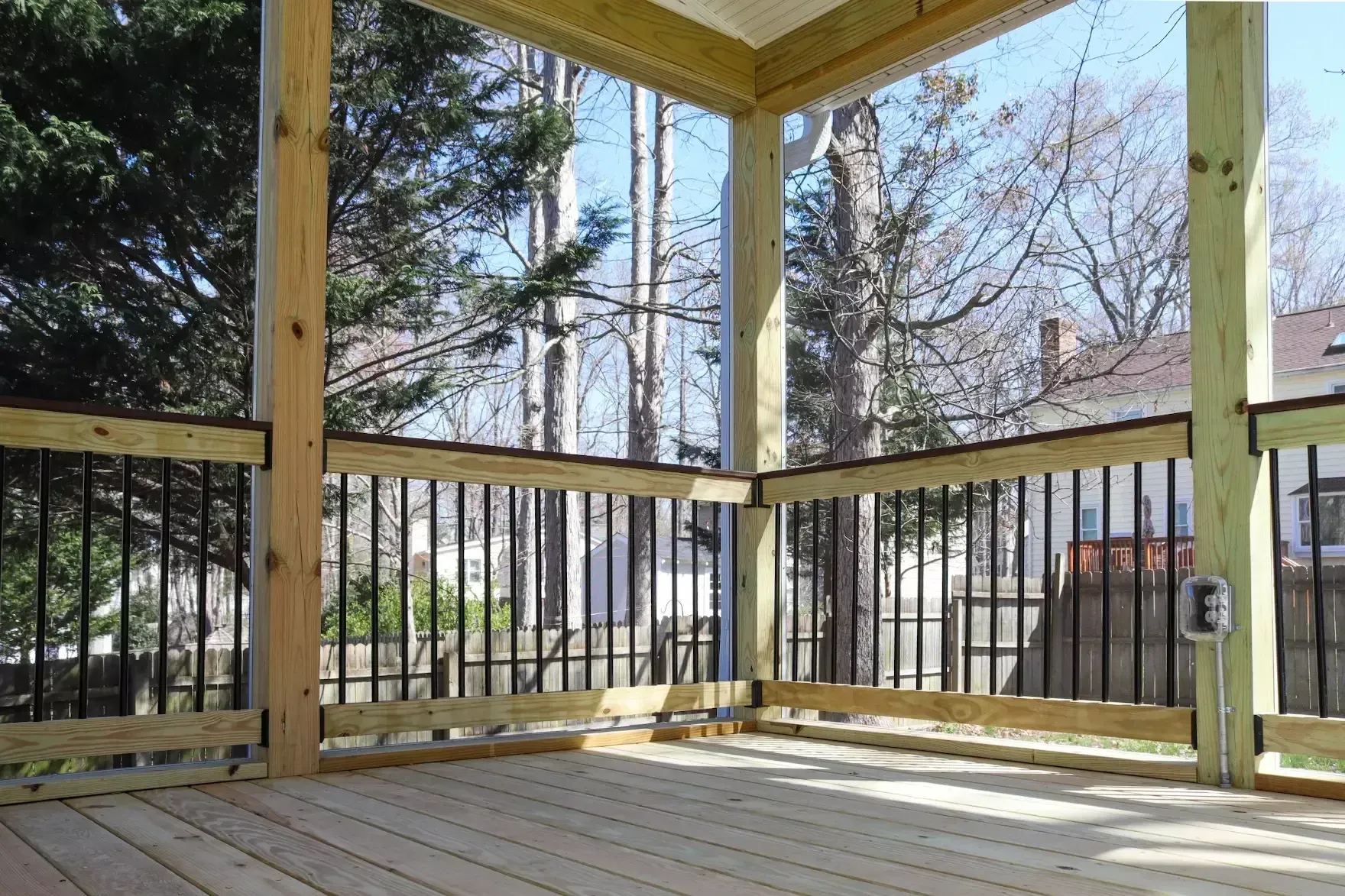 A wooden deck with black metal railings and vertical support posts, overlooking a backyard with trees and houses.