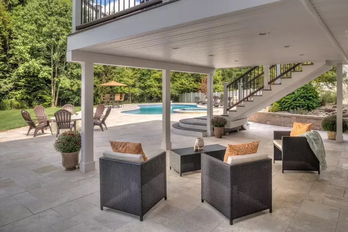 Covered patio under a raised deck, featuring outdoor chairs, a small table, and stairs leading to a backyard pool.
