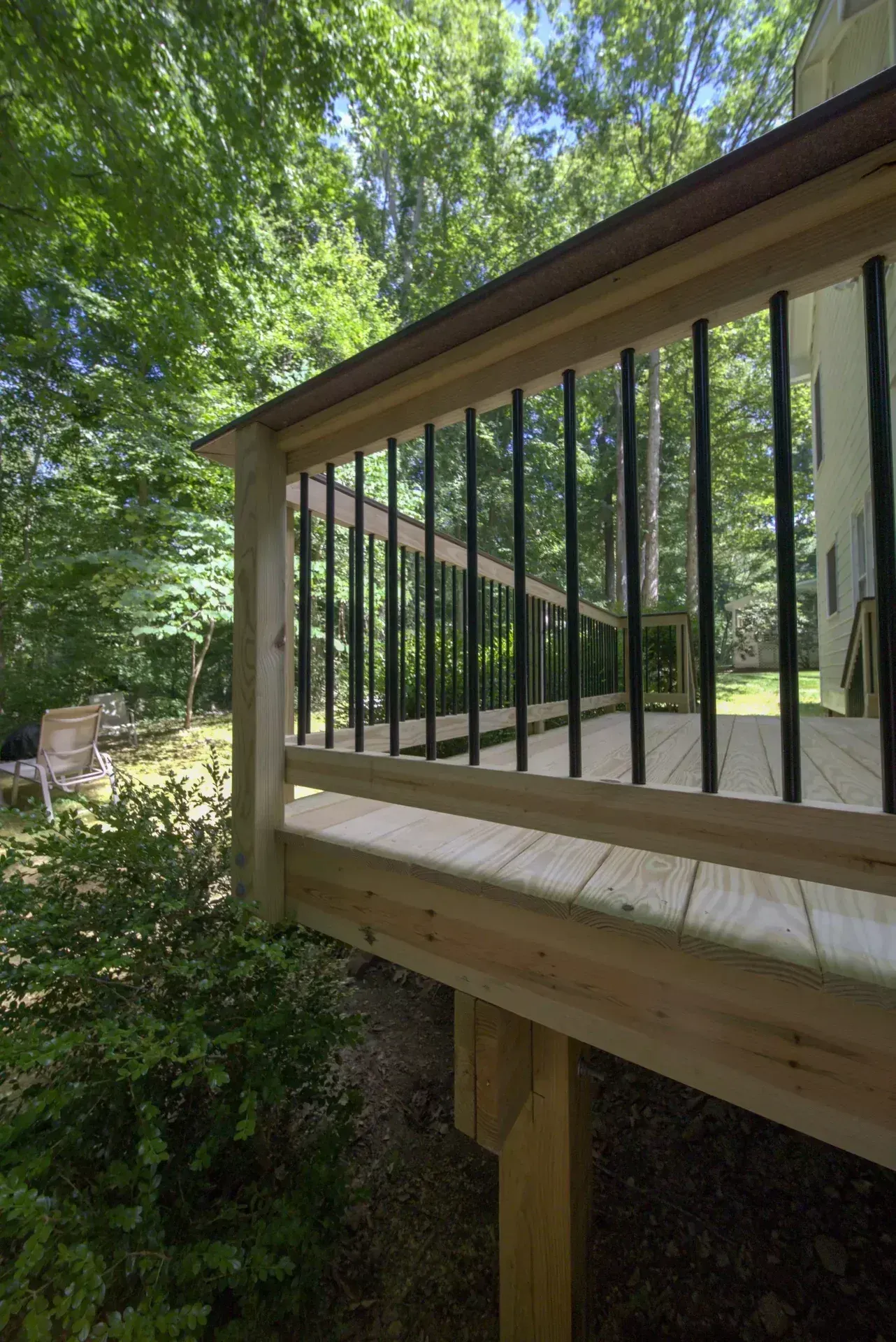 A view of a new wooden deck with black metal balusters, overlooking a lush, sunlit backyard forest.