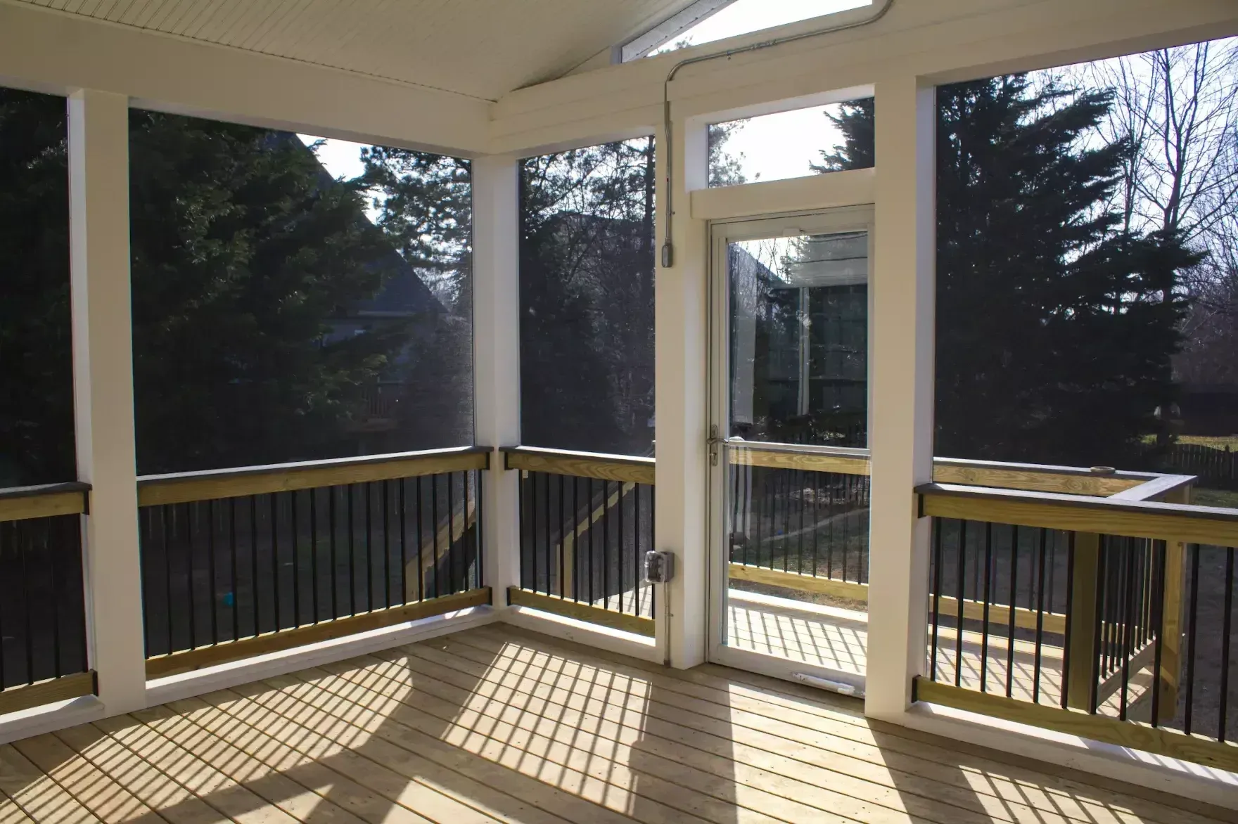 An outdoor screened-in porch featuring light wood decking, beige pillars, black railings, and a glass-paneled door.