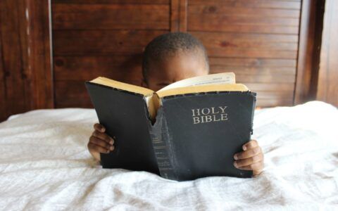 a young boy is reading a bible on a bed