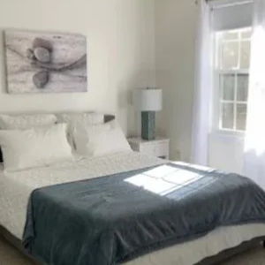 A bedroom with a queen-sized bed, white bedding, a grey throw blanket, two nightstands, and a window with white curtains.