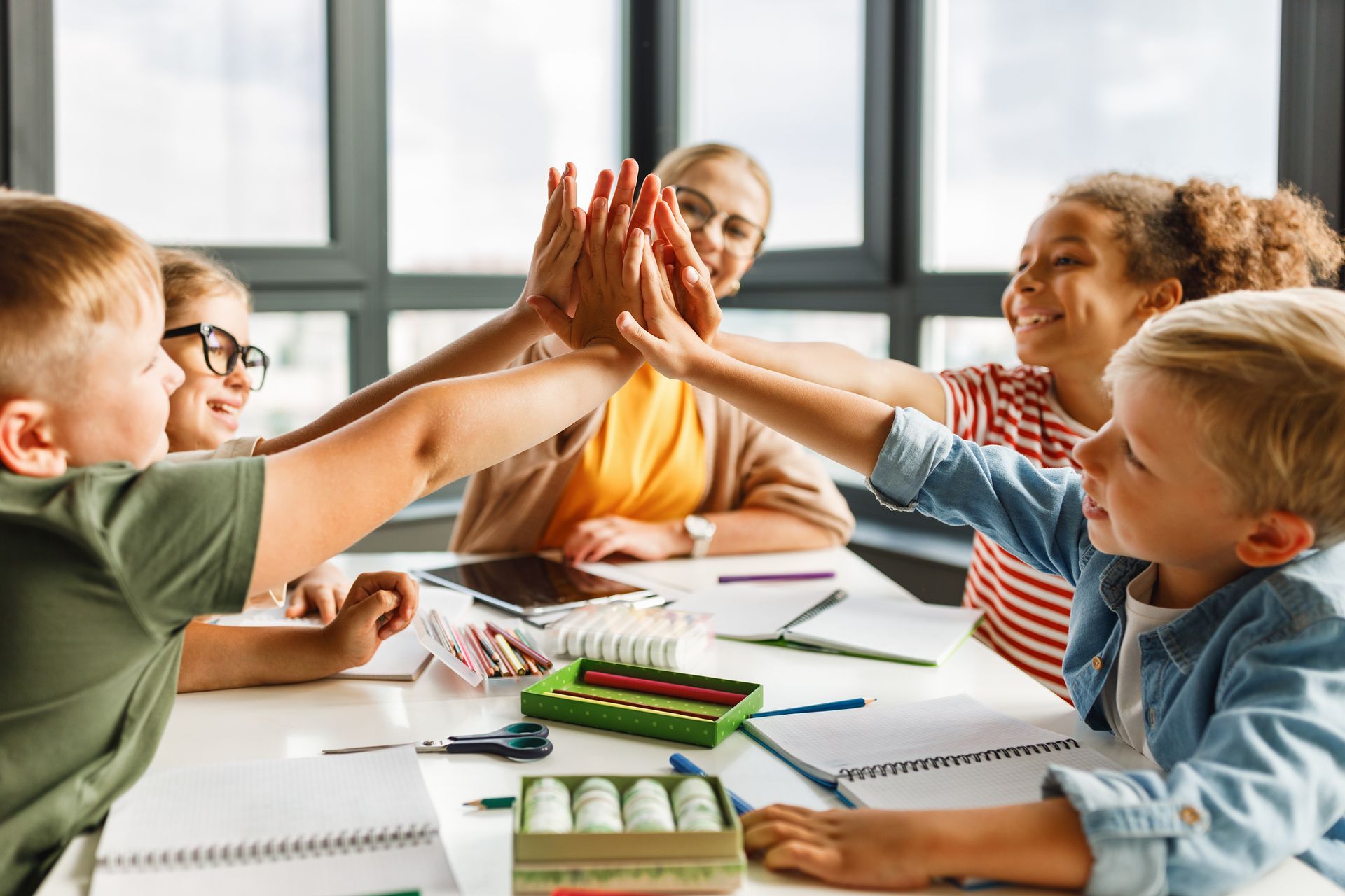 Children and a teacher high-five around a classroom table with art supplies and notebooks