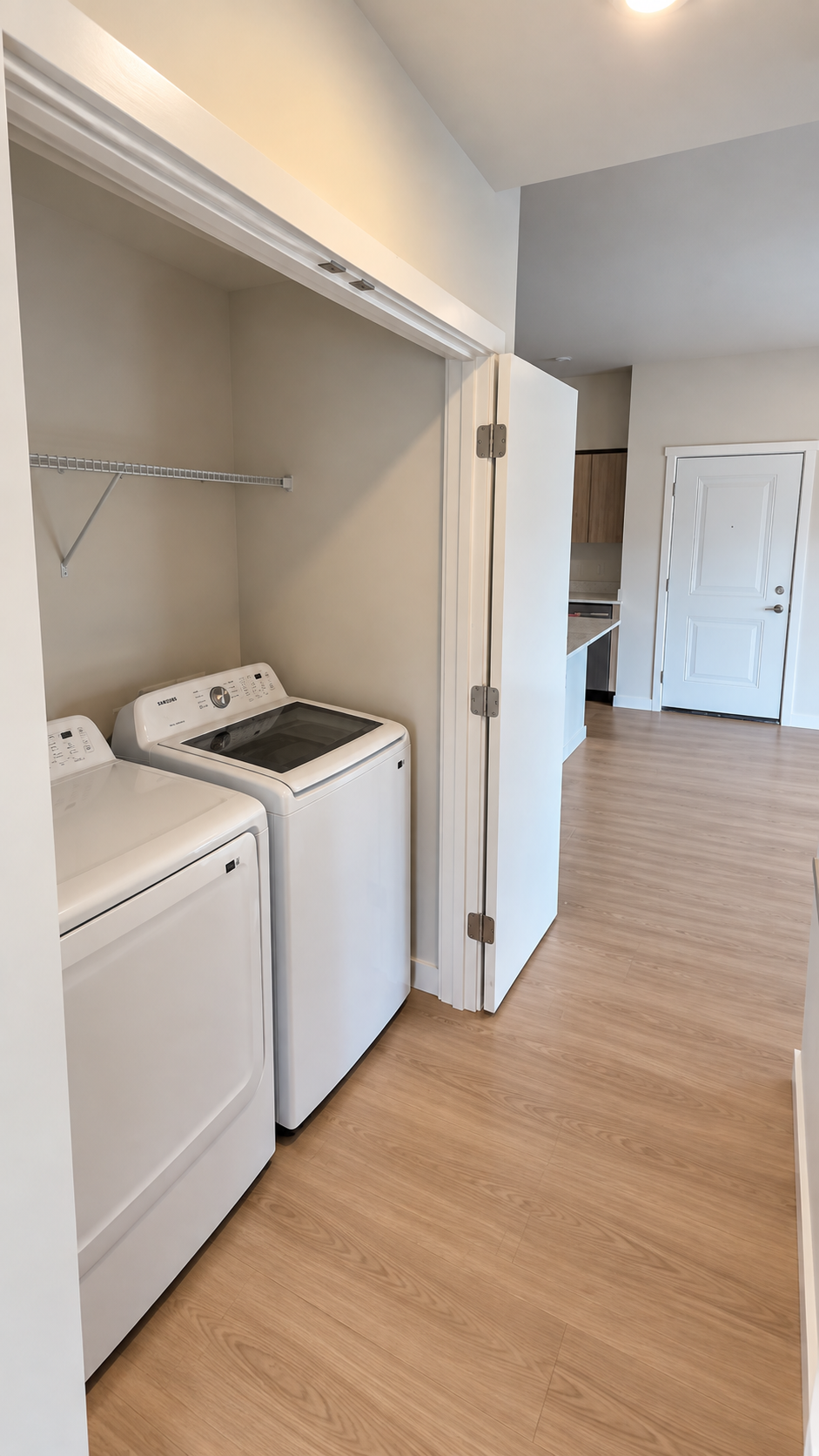 Laundry closet with a washer and dryer beside an open door in a wood-floored apartment hallway