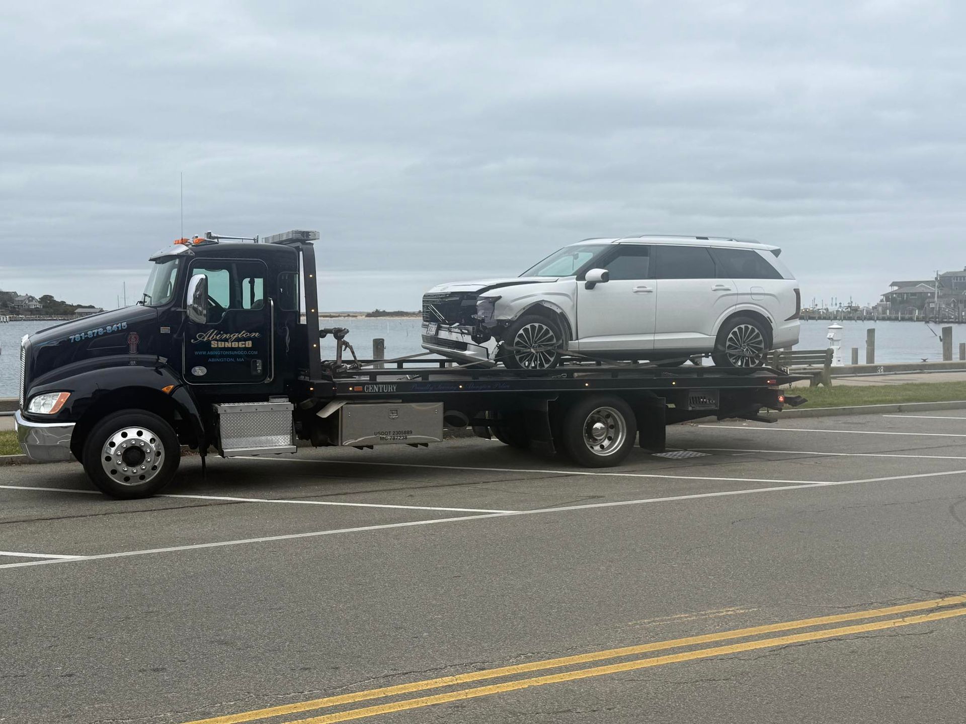 White SUV with front-end damage loaded onto a tow truck, parked by water.