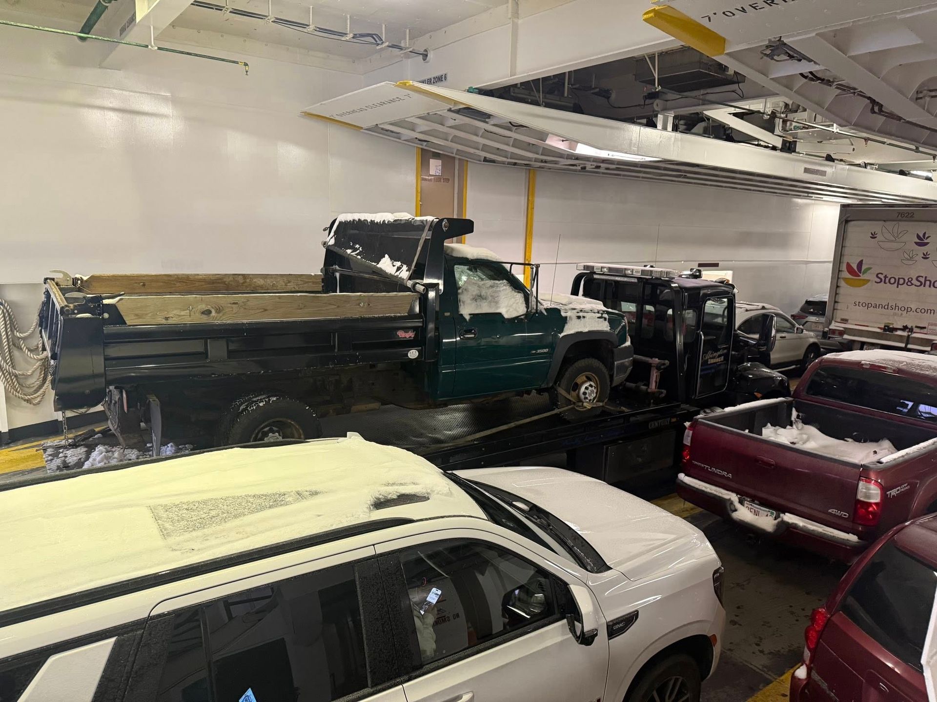 Vehicles, including a green dump truck, on a ferry, covered in snow. Interior shot.