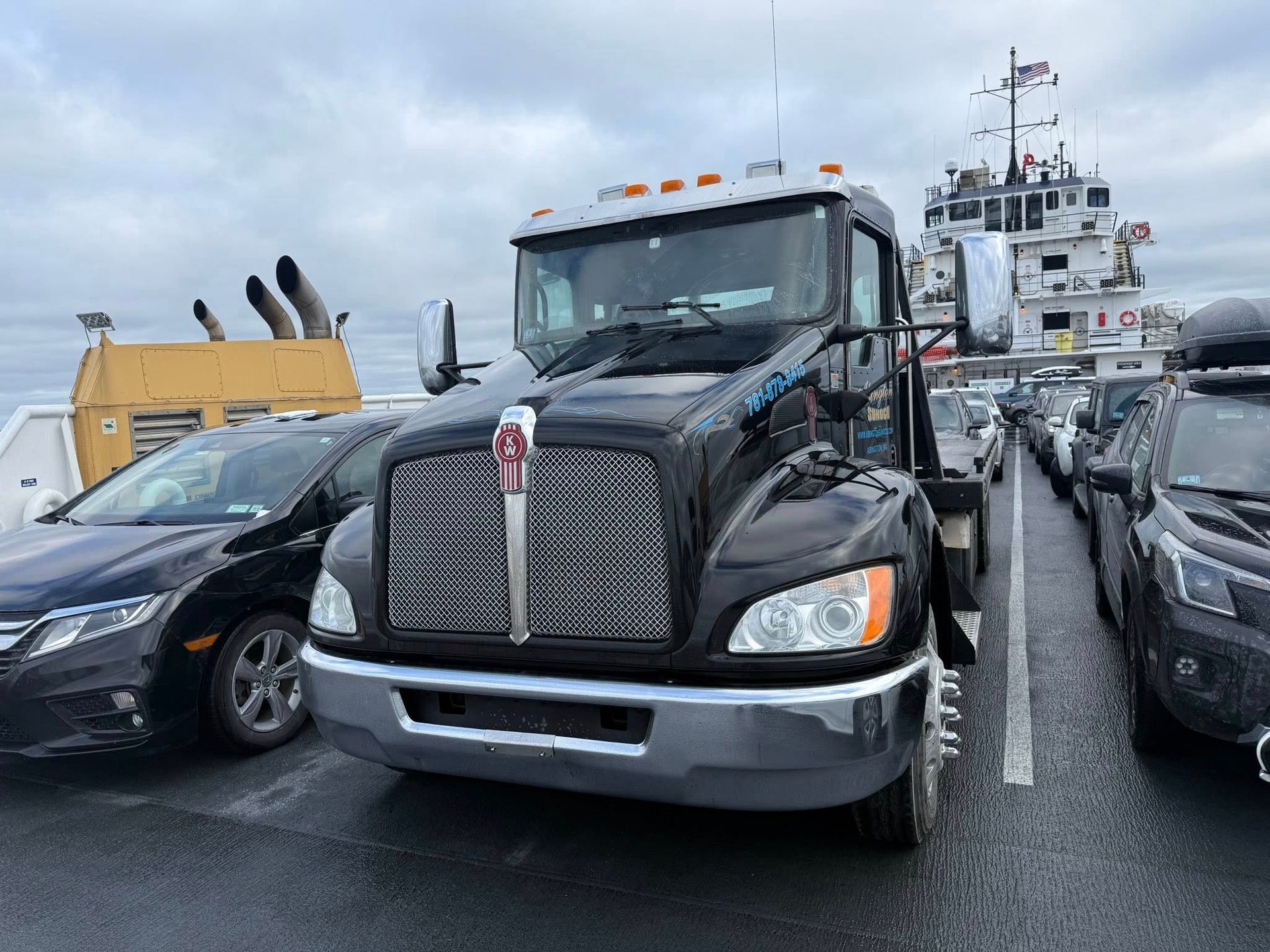 Black semi-truck parked on a ferry, surrounded by cars. Cloudy sky in the background.