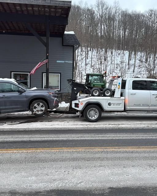 Tow truck towing a gray car, a small tractor is on the truck bed, snowy roadside, building backdrop.
