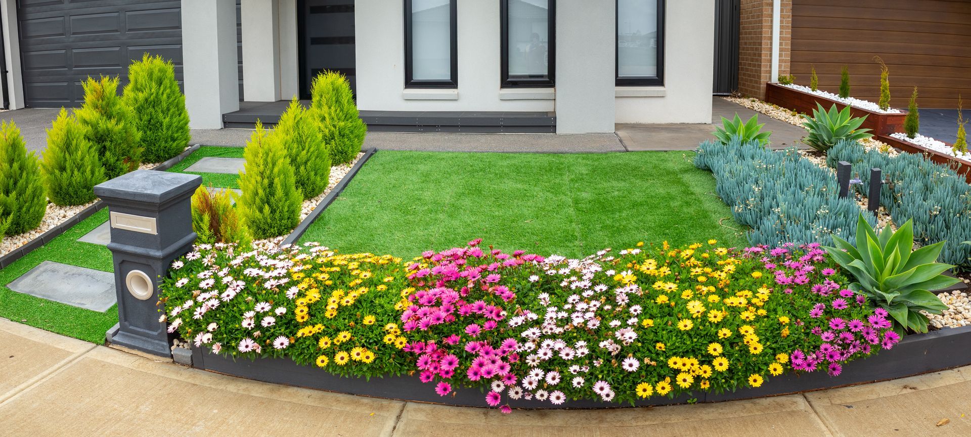 Front yard flower bed with pink, yellow, and white blooms edging a modern house walkway