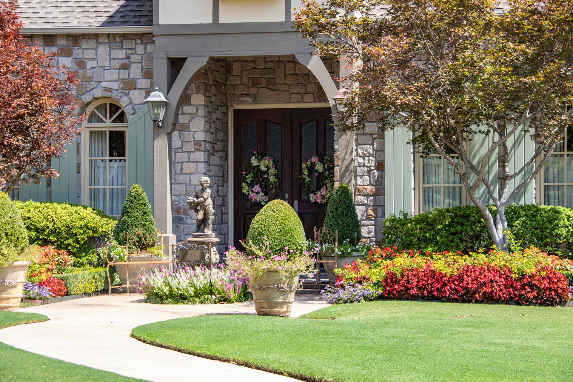 Stone house entryway with arched doorway, manicured lawn, colorful flower beds, and potted plants