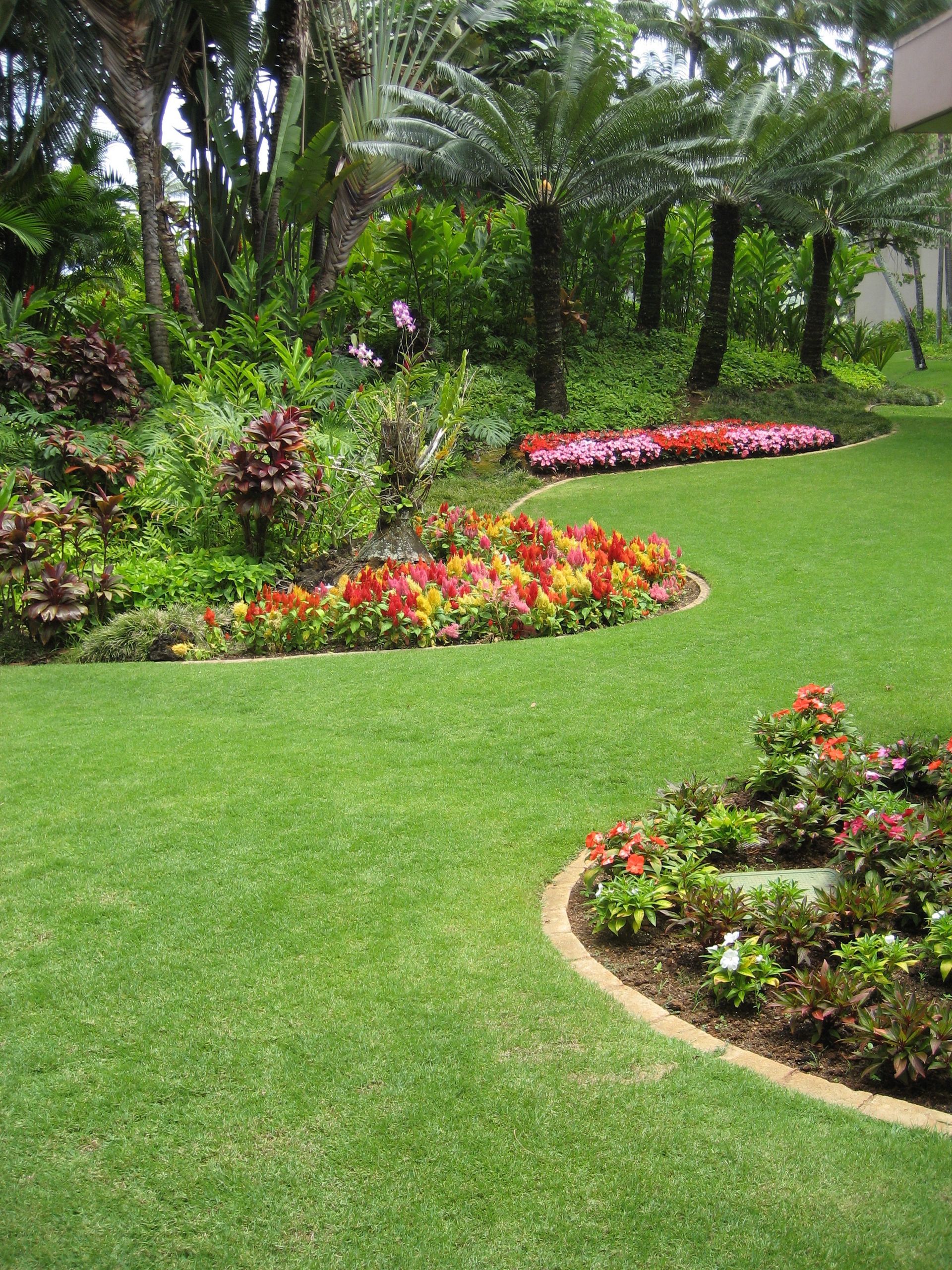 Lush garden with green lawn, colorful flower beds, and tall trees in bright sunlight