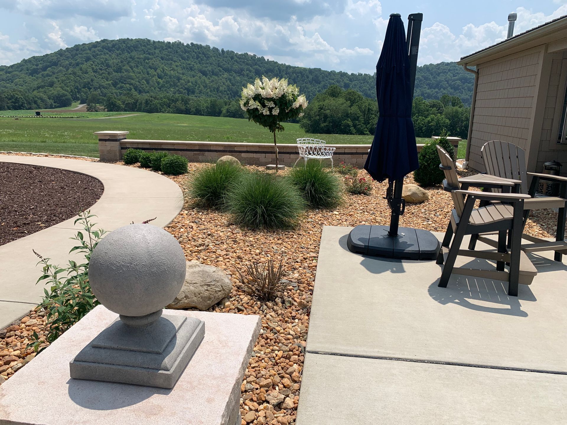 Patio with table, closed umbrella, stone sphere, and landscaped yard with hills in the background