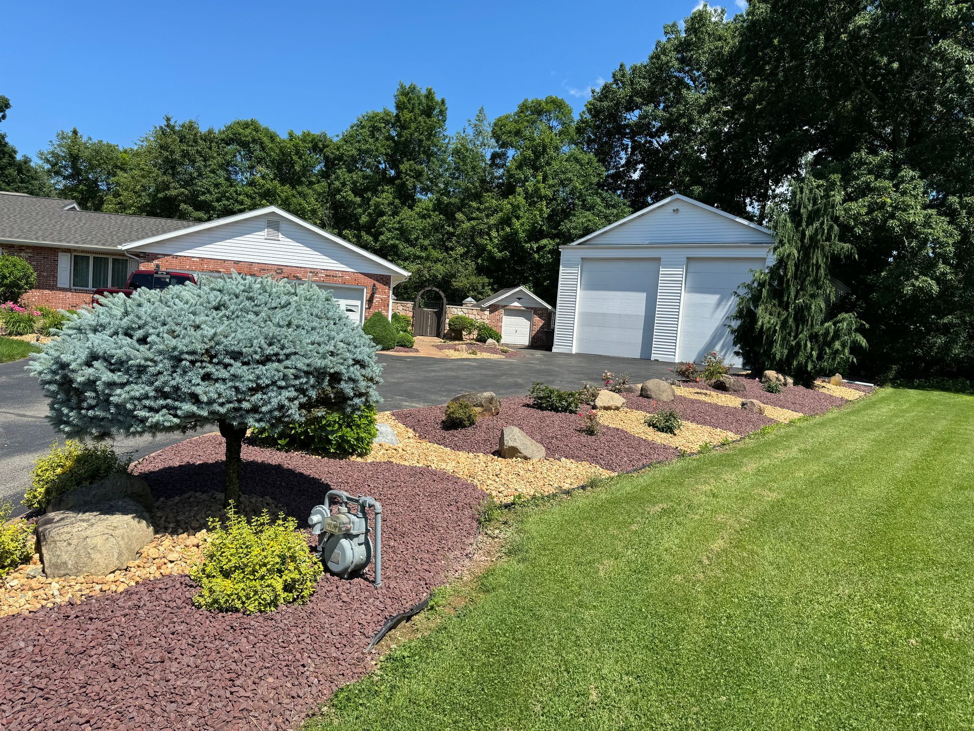Landscaped front yard with mulch beds, shrubs, and a white shed beside a green lawn under a blue sky