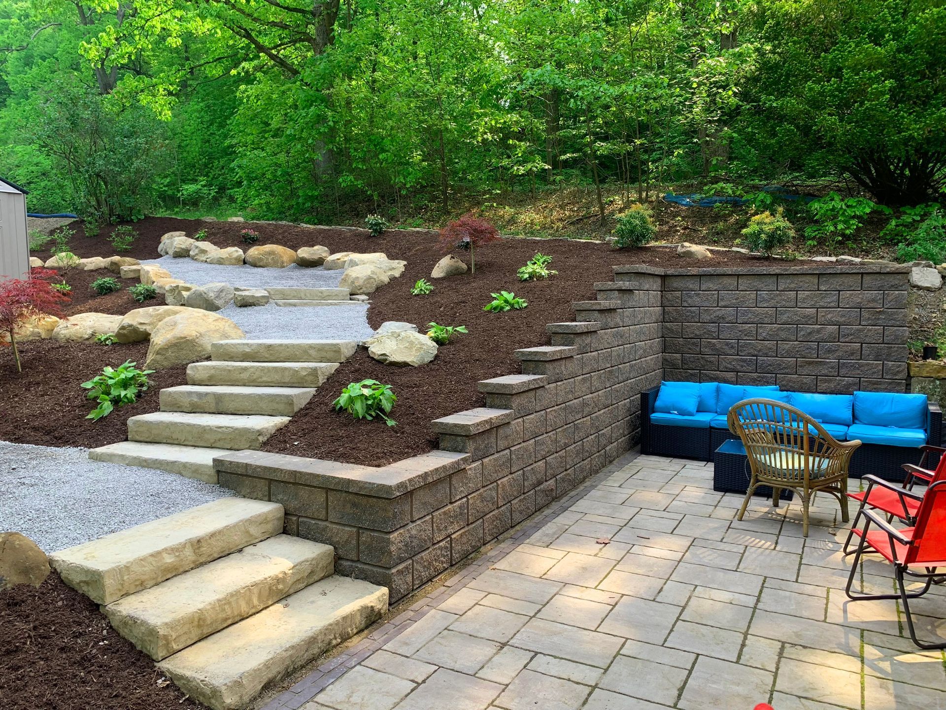 Stone patio with steps, retaining walls, and a blue outdoor sofa in a landscaped backyard.