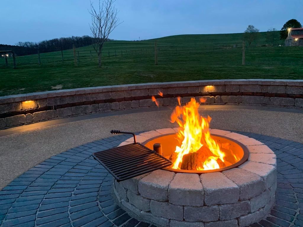 Stone fire pit with a bright flame in a curved brick patio at dusk, overlooking a grassy hillside