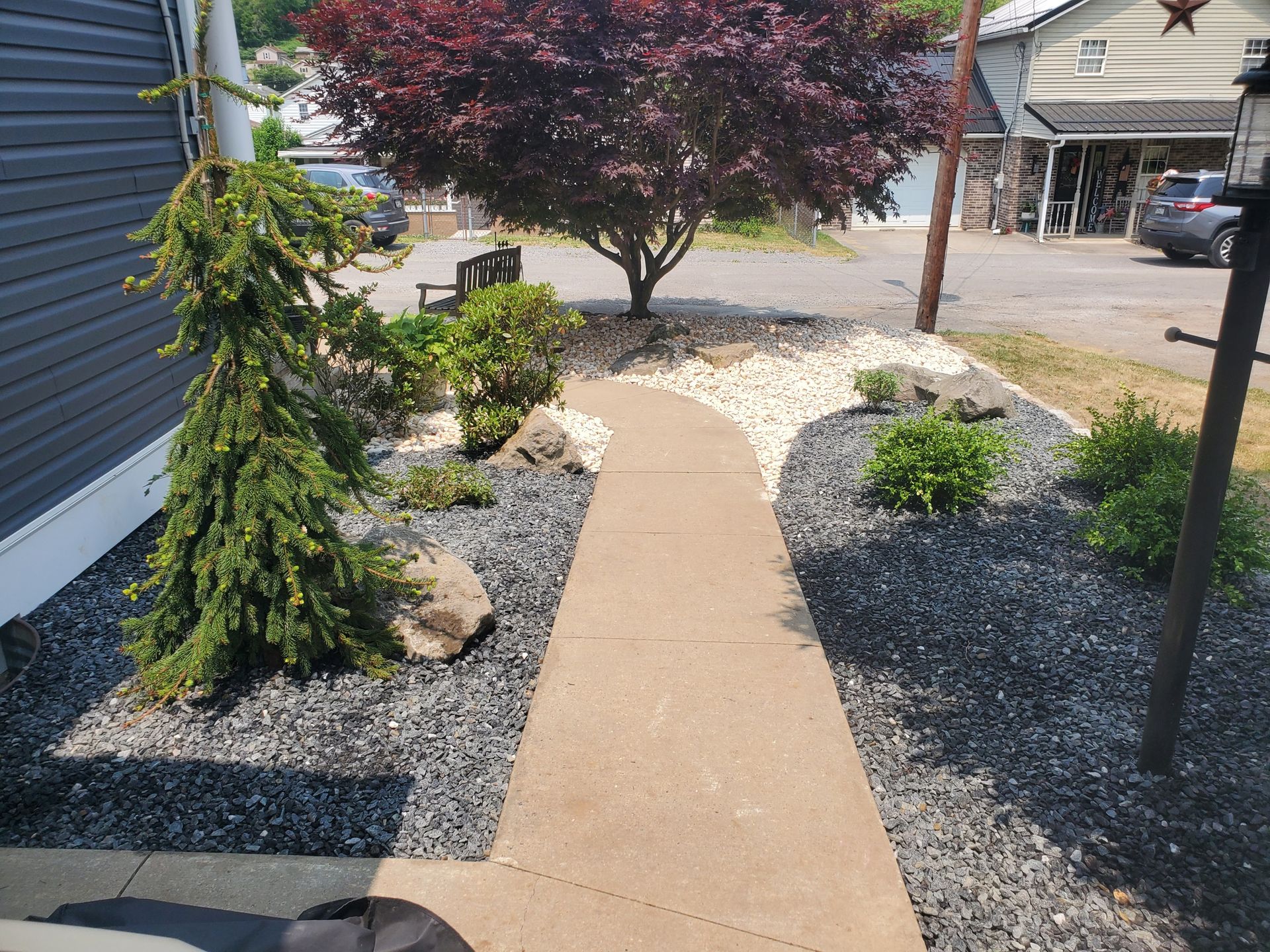 Curved sidewalk through a gravel landscaped yard with shrubs and a red-leaf tree beside buildings
